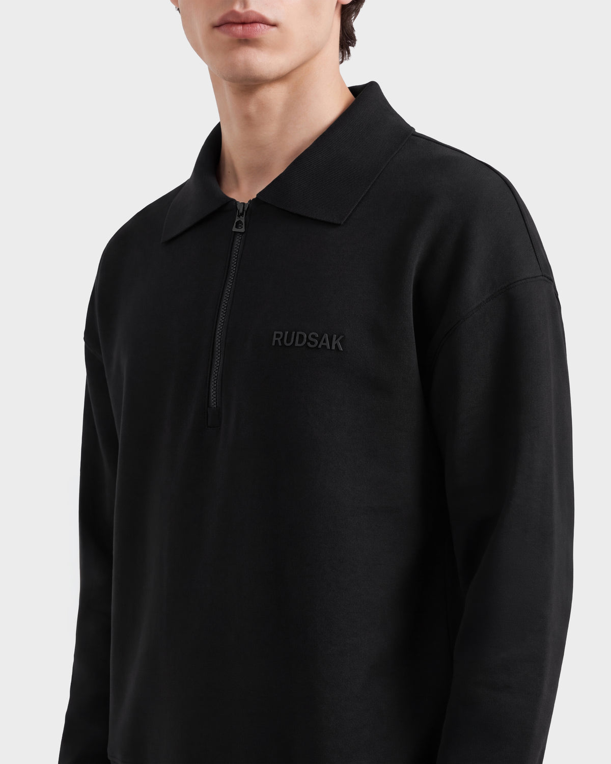 Someone wears the IDRIS - 6526500 Black, a black long-sleeve half-zip polo collar sweater by Rudsak with “RUDSAK” embroidered on the chest, against a plain light background.