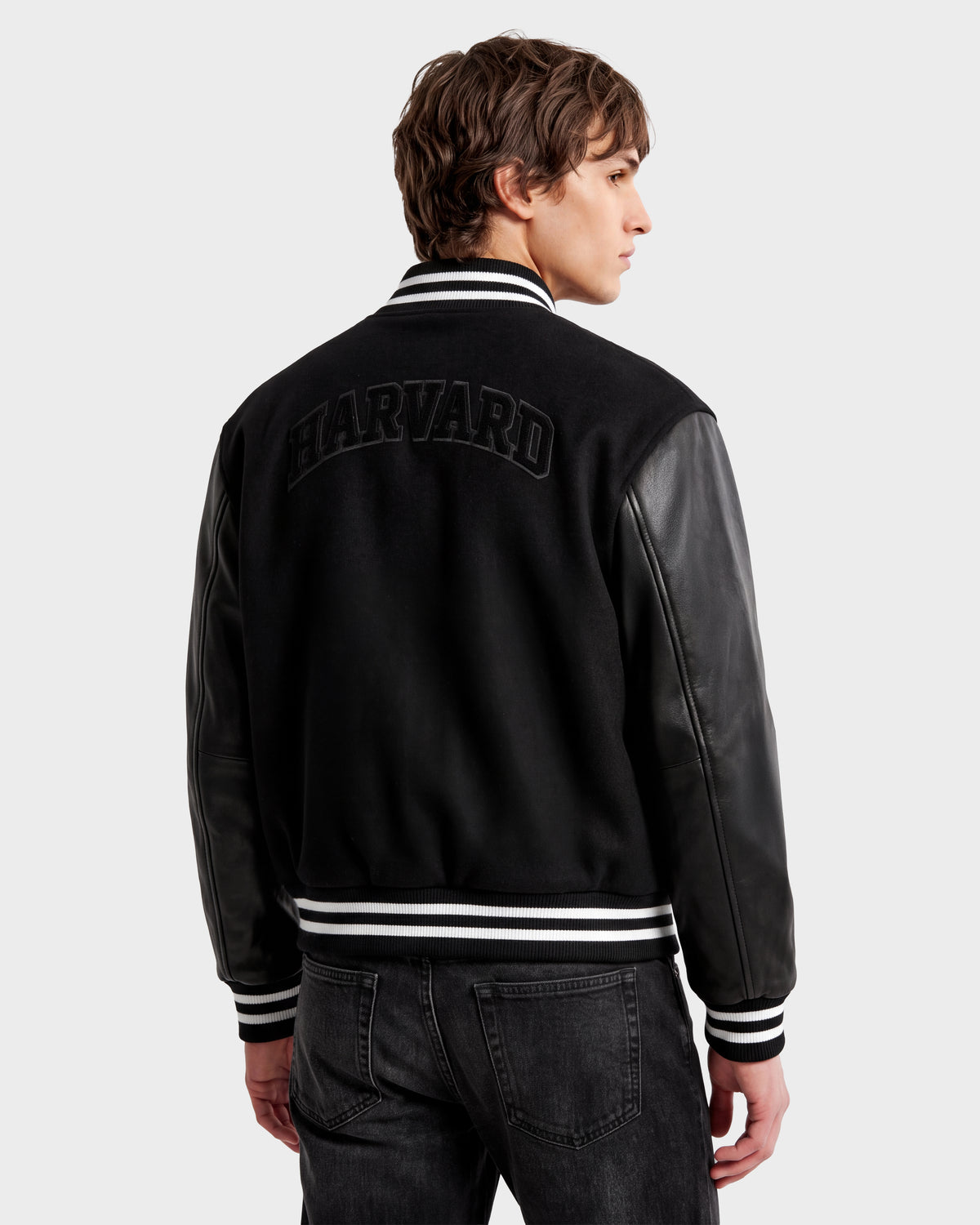 A person wearing the Rudsak JOHN HV - H125504 Black varsity jacket with leather sleeves and “HARVARD” boldly emblazoned on the back stands facing away, looking to the side—a timeless nod to limited edition collegiate jacket style.