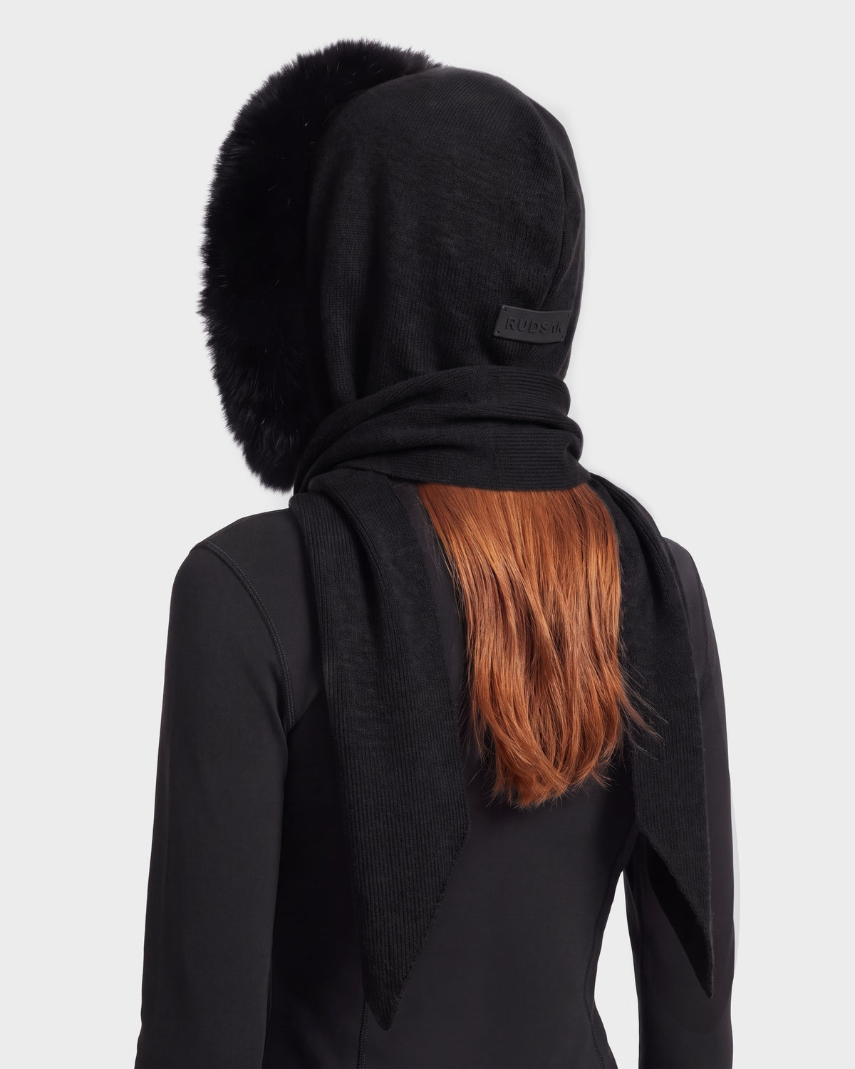 A person with long reddish hair is seen from behind wearing the KARYMA - 8426507 Black hood-and-scarf hybrid by Rudsak, featuring plush trim over a black long-sleeve top. The accessory includes a label on the back and adds a stylish touch to any winter ensemble, set against a light gray background.