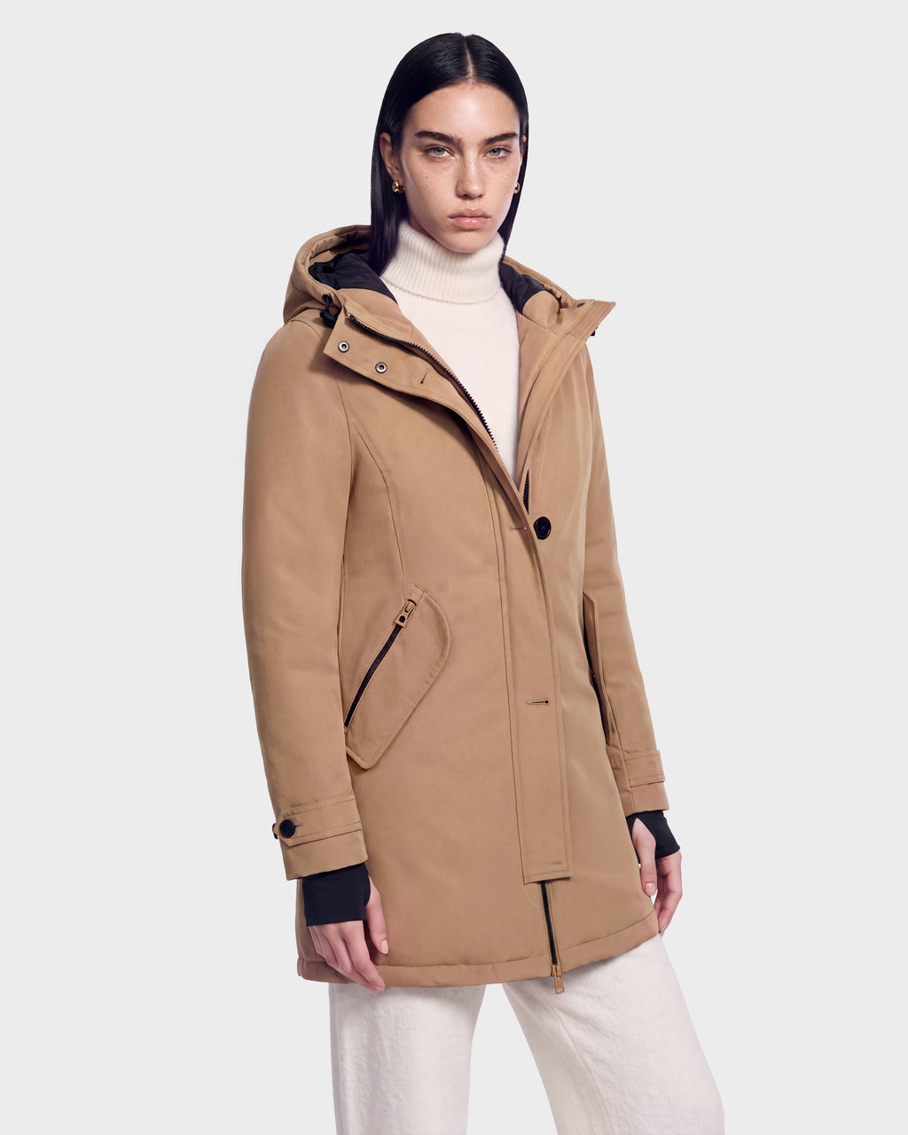 A woman with straight dark hair wears the KEYSHIA - 8126517 Camel by Rudsak over a cream turtleneck and white pants, standing against a plain light gray background.