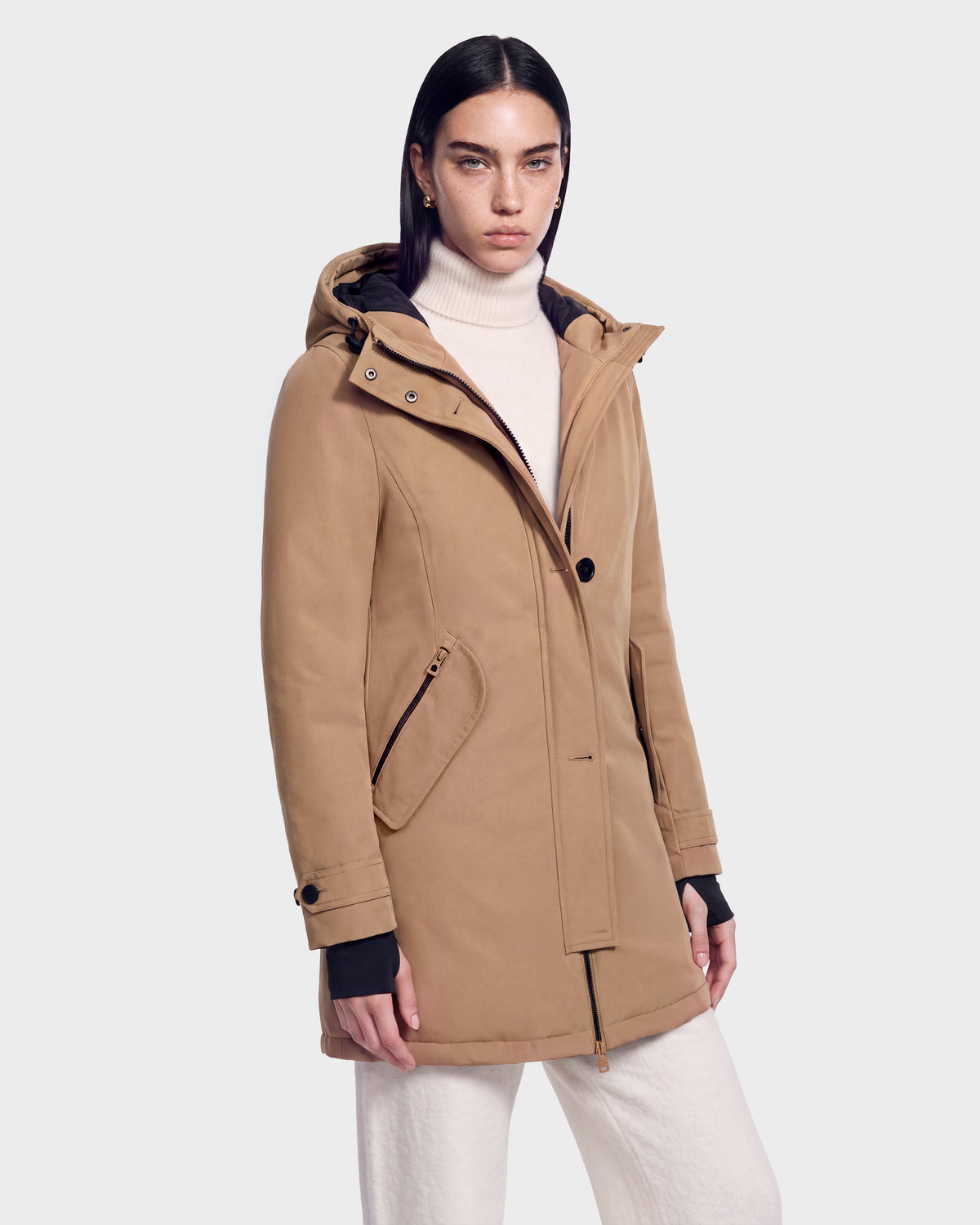 A woman with straight dark hair wears the KEYSHIA - 8126517 Camel by Rudsak over a cream turtleneck and white pants, standing against a plain light gray background.