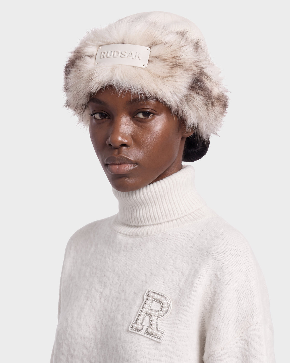 A person wearing a faux fur trapper hat and a LORETTA W - 8426510 Vanilla cream turtleneck sweater with an embellished letter R patch.