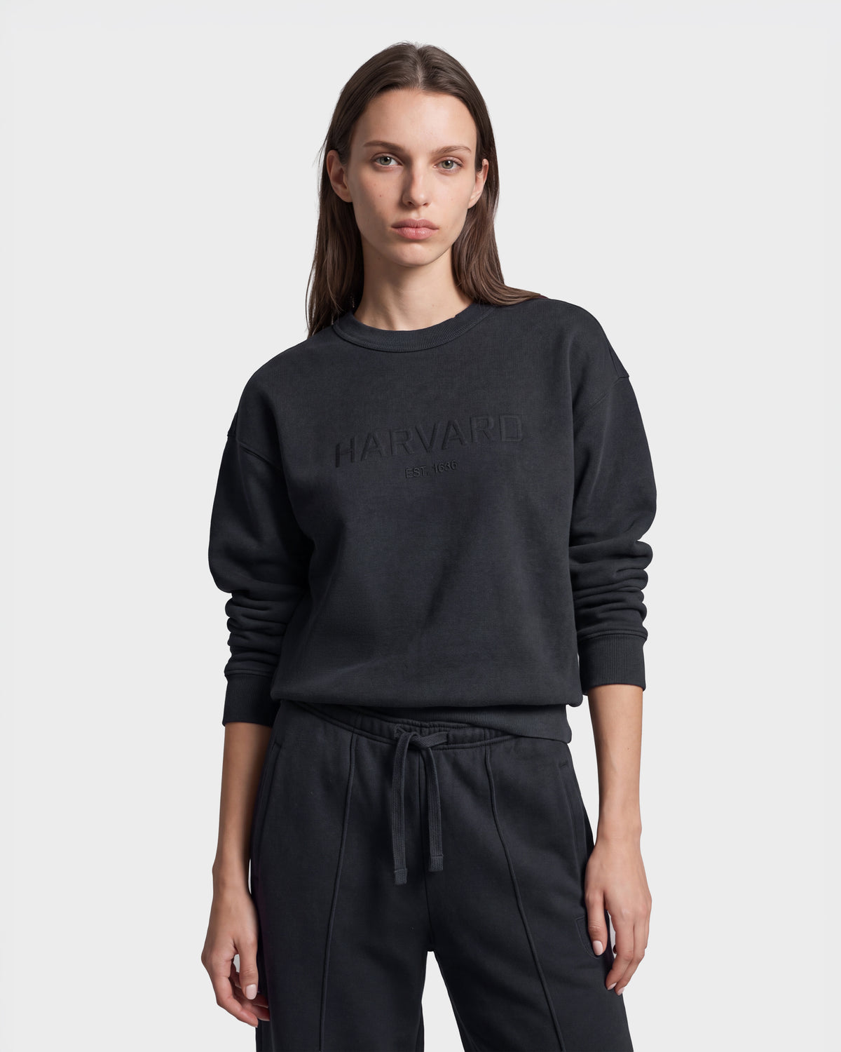 A woman with long brown hair models the Rudsak LEXYE HV - H526900 Titanium sweatshirt and matching black sweatpants, standing against a plain light gray background.