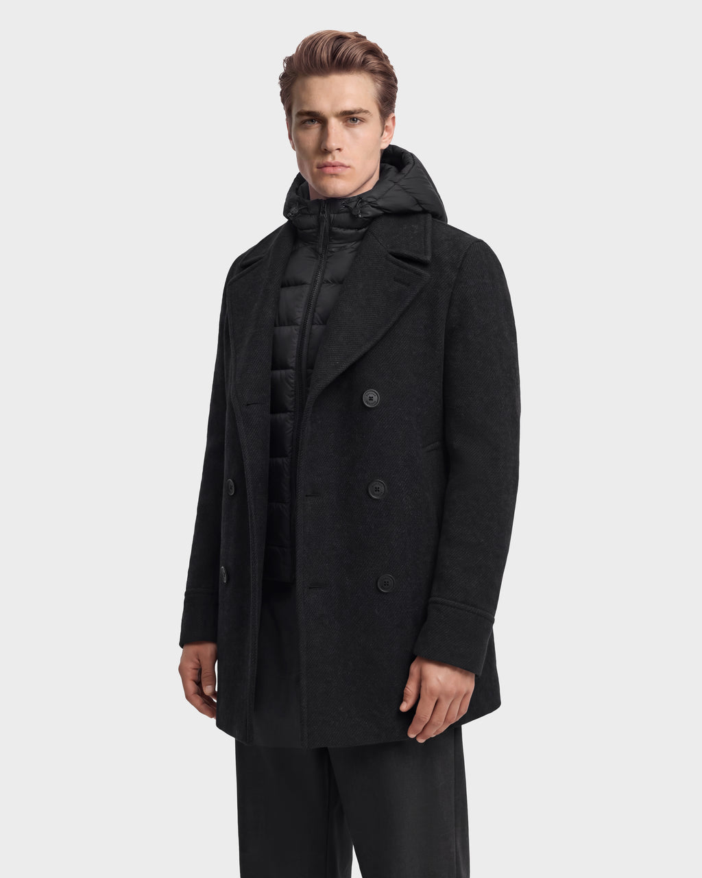 A man wearing a charcoal LUCIEN - 6126508 Rudsak double-breasted wool-blend coat over a black quilted hooded vest with duck down insulation.