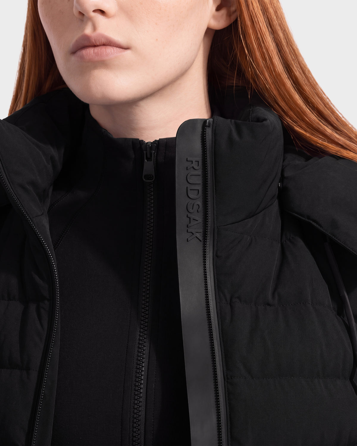 A woman with long red hair wears the Rudsak MERINA - 8125056 Black quilted down jacket, partially unzipped to reveal a zip-up top underneath. This lightweight transitional piece features the brand name RUDSAK embossed on a vertical leather strip near the zipper.