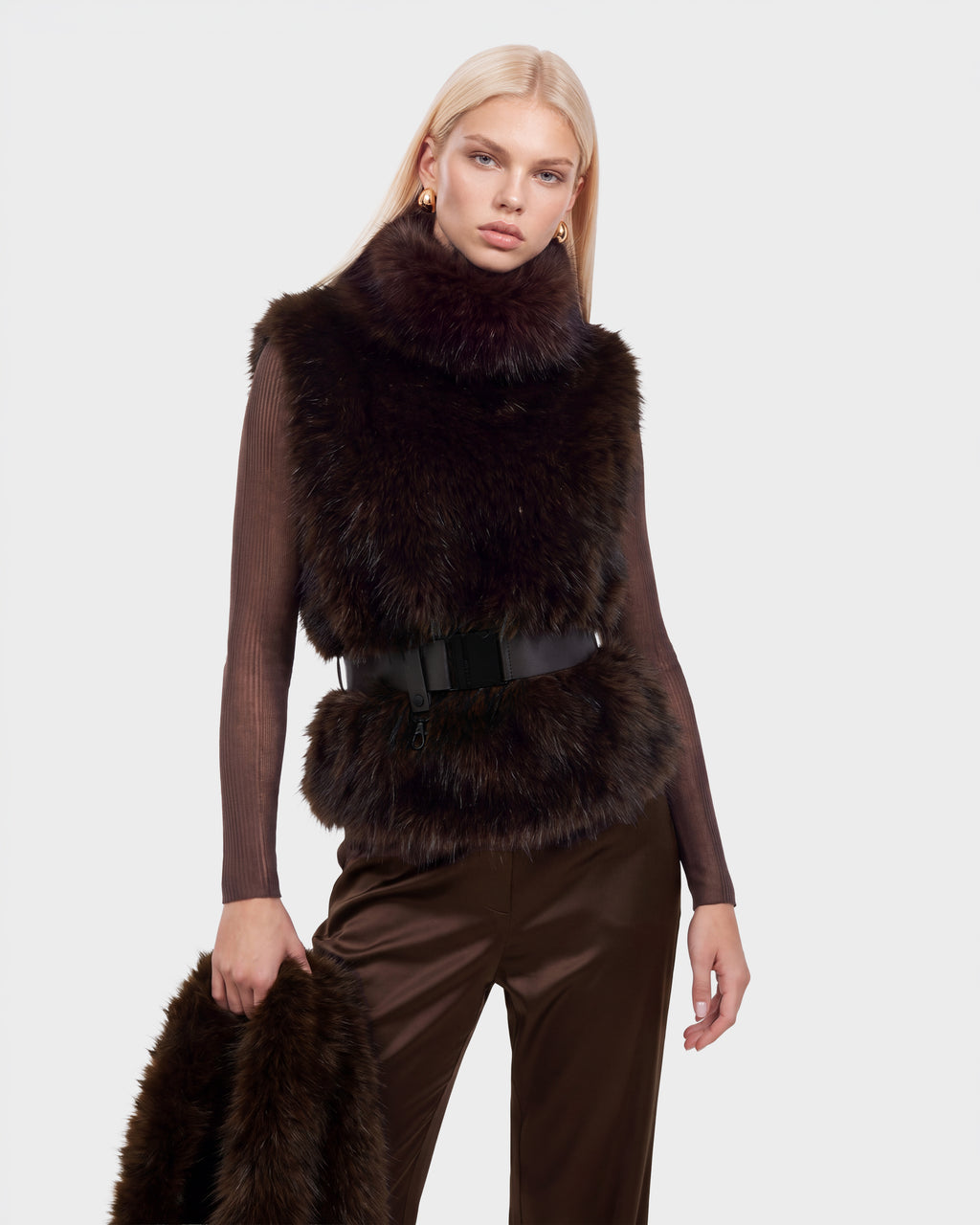 A person wears a MAULIN - 8524611 Mix Espresso brown faux fur vest over a long-sleeved shirt and trousers, cinched with a wide black belt, while holding a matching fur accessory.