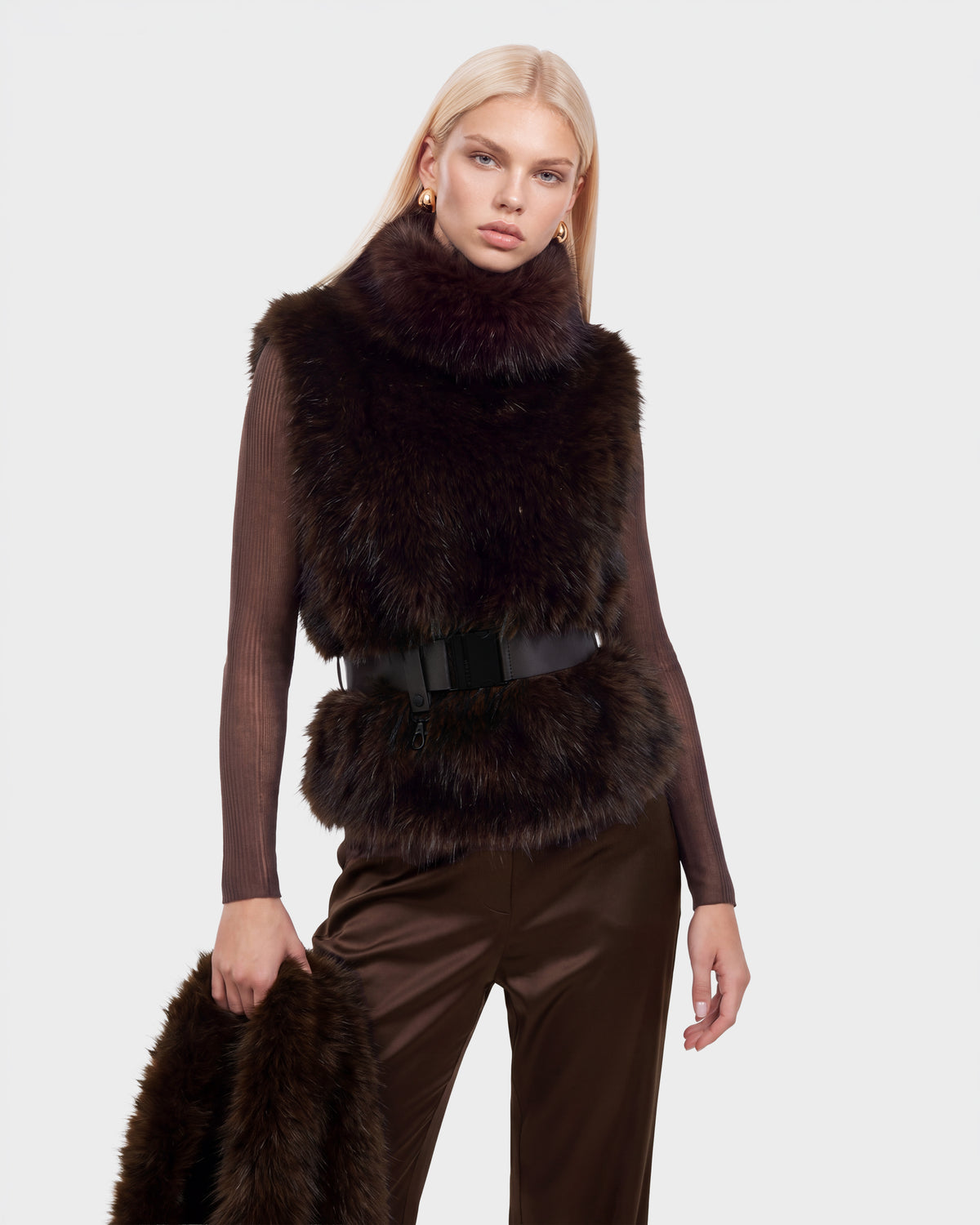 A person wears a MAULIN - 8524611 Mix Espresso brown faux fur vest over a long-sleeved shirt and trousers, cinched with a wide black belt, while holding a matching fur accessory.