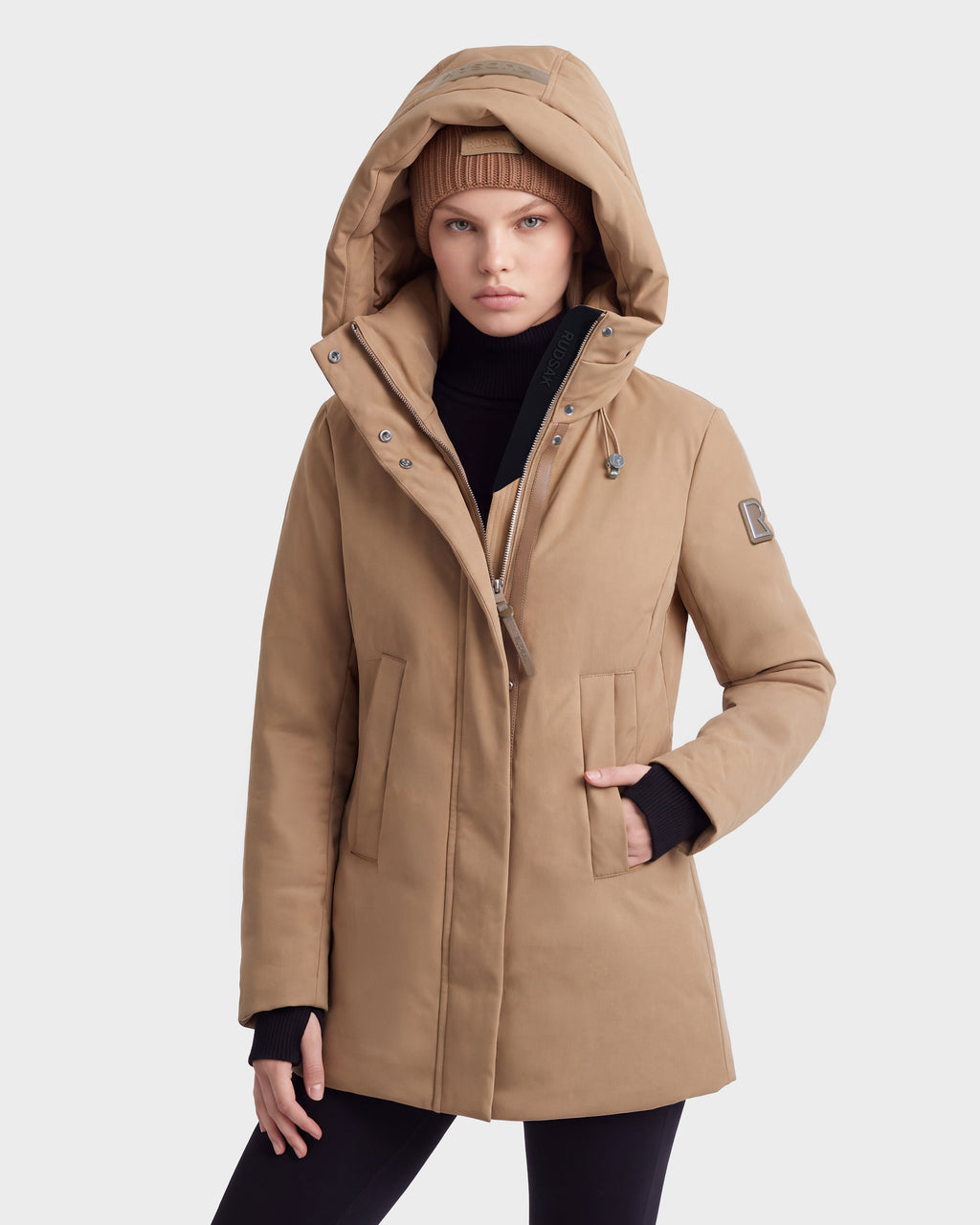 A person wears the Rudsak OPAL - 8126618 Camel parka with RDS-certified down, a tan beanie, and black turtleneck, standing against a plain light background with one hand in their pocket.