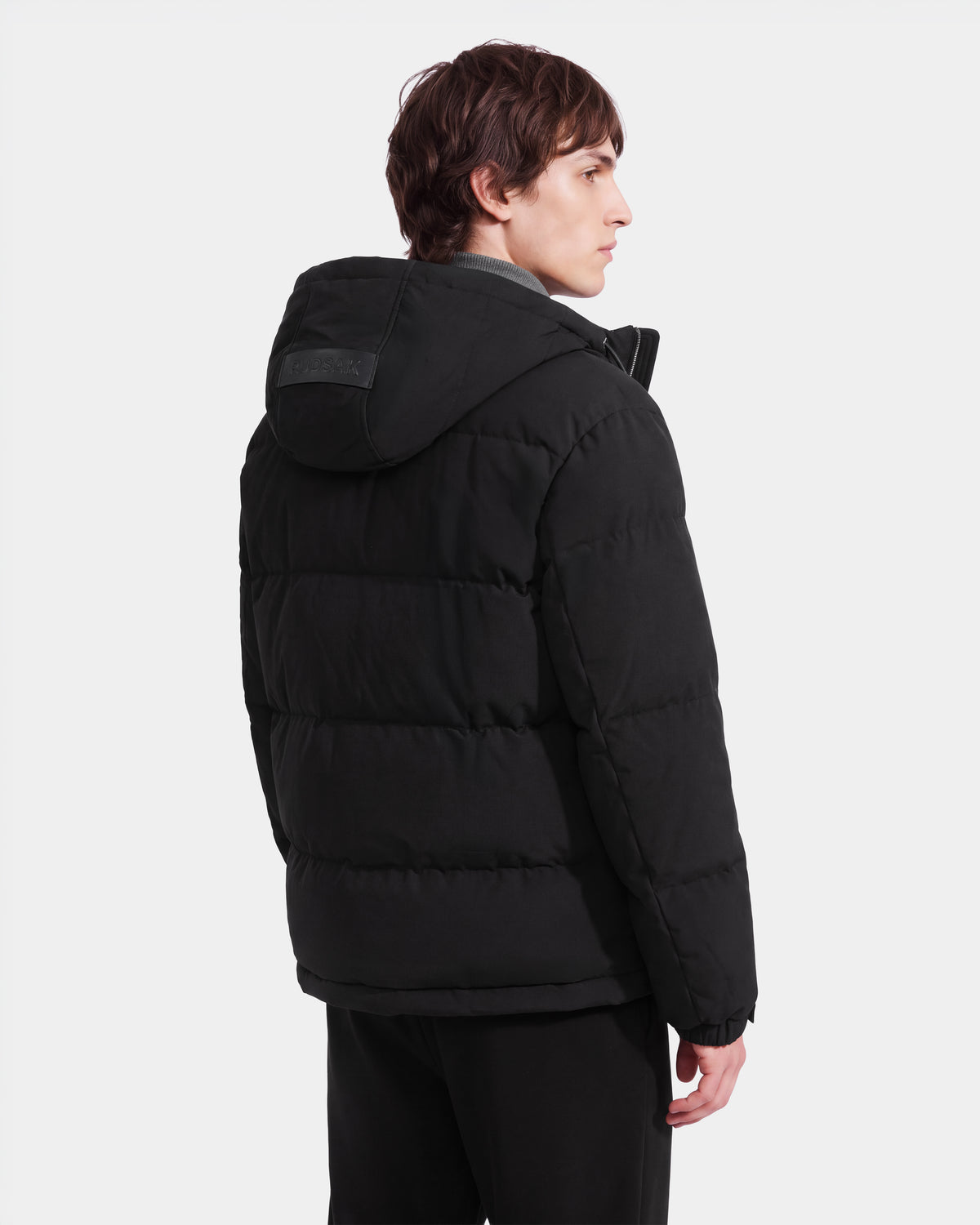 A person with short brown hair is standing and facing away, displaying the back and left side of the Rudsak STERLING EL - 6126759 Black puffer jacket with a hood against a plain white background.