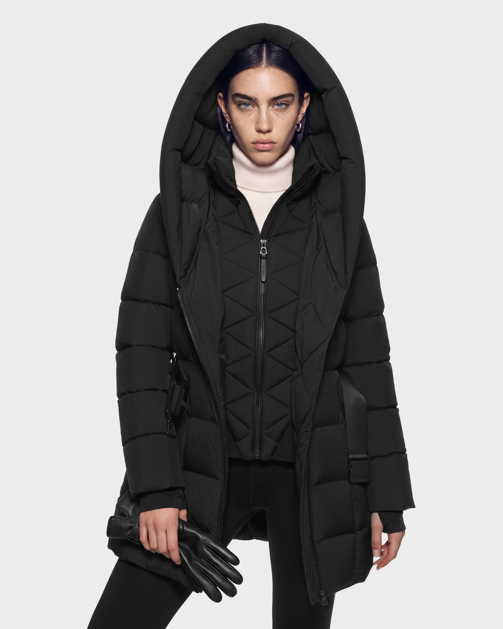 A person wearing a black Rudsak TAYLOR M - 8125965 Black puffer coat with a large hood over a white turtleneck, holding a pair of black gloves.