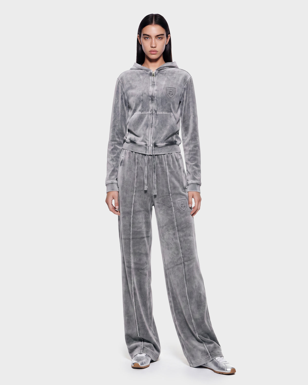 A person stands facing forward against a plain light background, wearing the Rudsak TYRA HV - H526904 Titanium gray velour tracksuit with a zip-up hoodie and wide-leg pants, paired with silver sneakers.