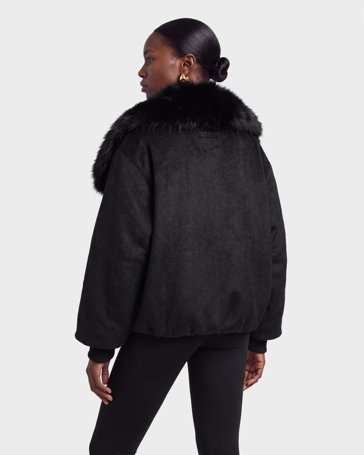 A woman stands facing away, wearing the Rudsak ZARIA - 8126999 Black jacket with a faux fur collar and black pants against a plain white background.