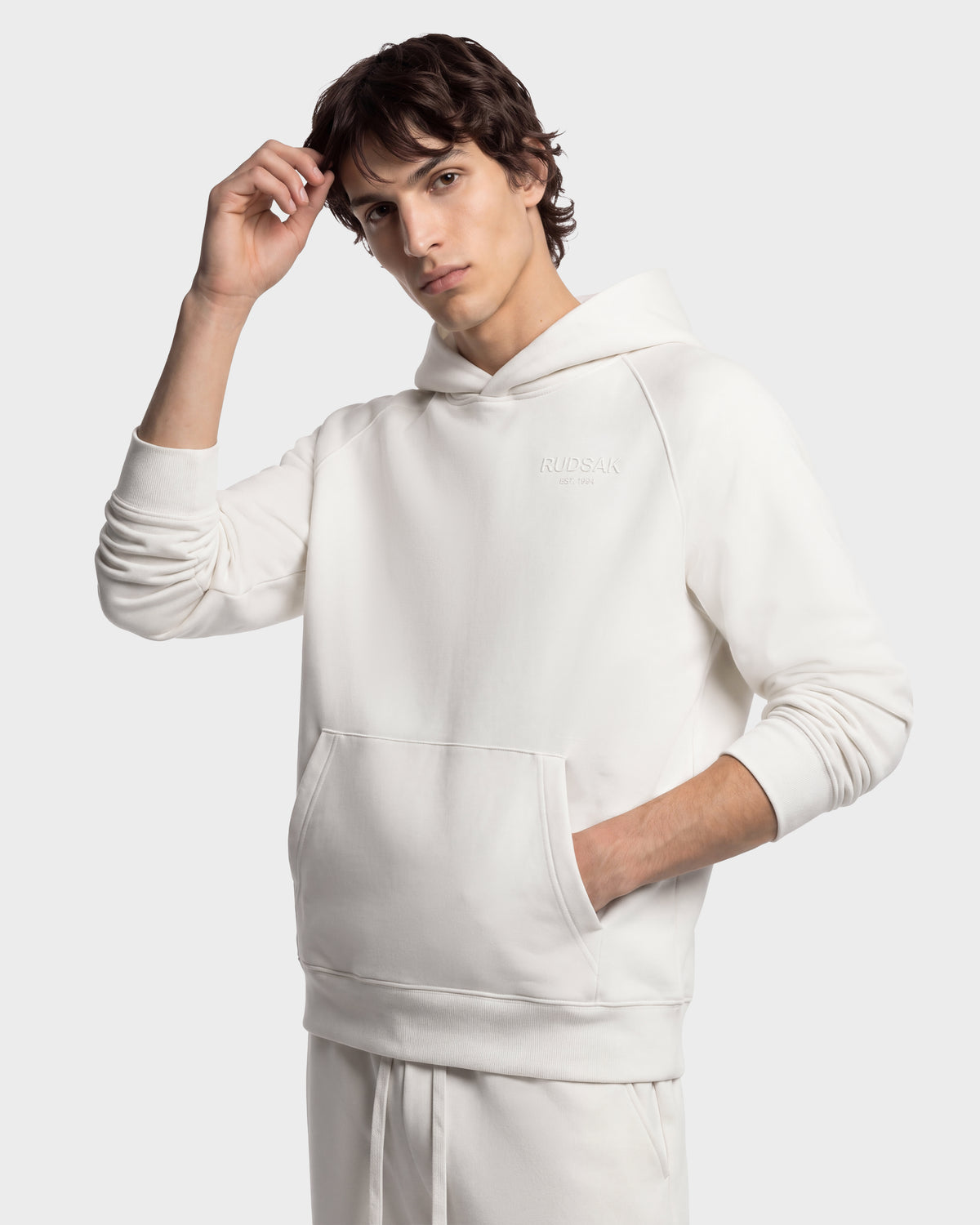 A person wearing a cream-colored Rudsak BRIAR - 9525001 Vanilla M unisex hooded sweatshirt, featuring a premium cotton blend, a front kangaroo pocket, and a small logo on the chest.