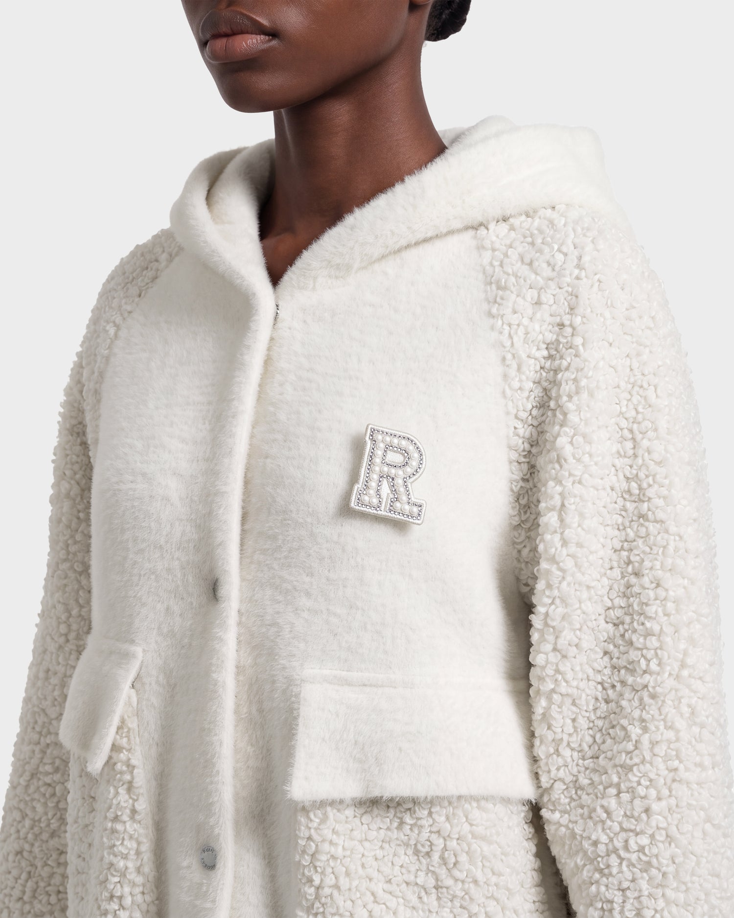 A person is shown from the shoulders up wearing the Rudsak DAKOTA - 8526515 Iceland textured white hooded coat, featuring large front pockets, snap buttons, and a bejeweled letter R pin on the chest.