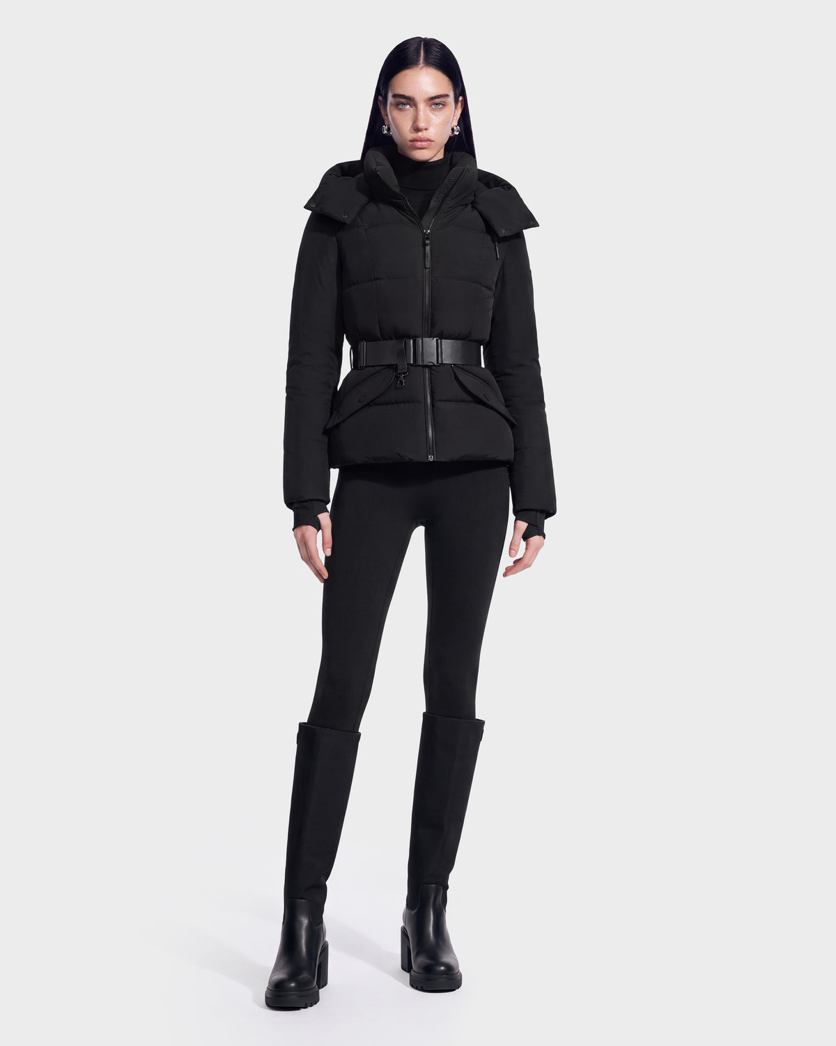 A person wears the ADELINE - 8124544 Black RDS-certified down puffer jacket by Rudsak, featuring a belt, paired with black pants and black knee-high boots, standing against a plain light background.