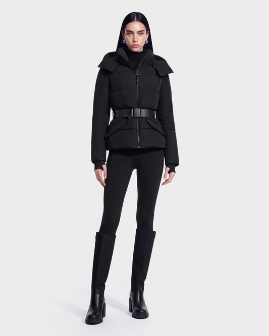 A person wears the ADELINE - 8124544 Black RDS-certified down puffer jacket by Rudsak, featuring a belt, paired with black pants and black knee-high boots, standing against a plain light background.