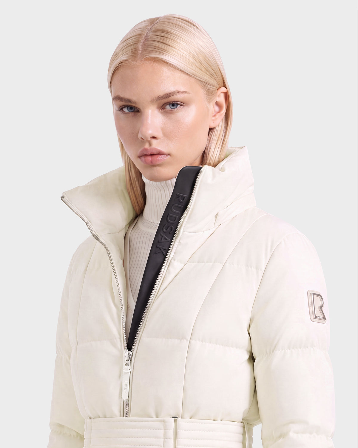 A woman wears a knee-length, vanilla-colored RUDSAK AUDREY L - 8124585 Vanilla belted puffer jacket with a high collar over a white turtleneck sweater.