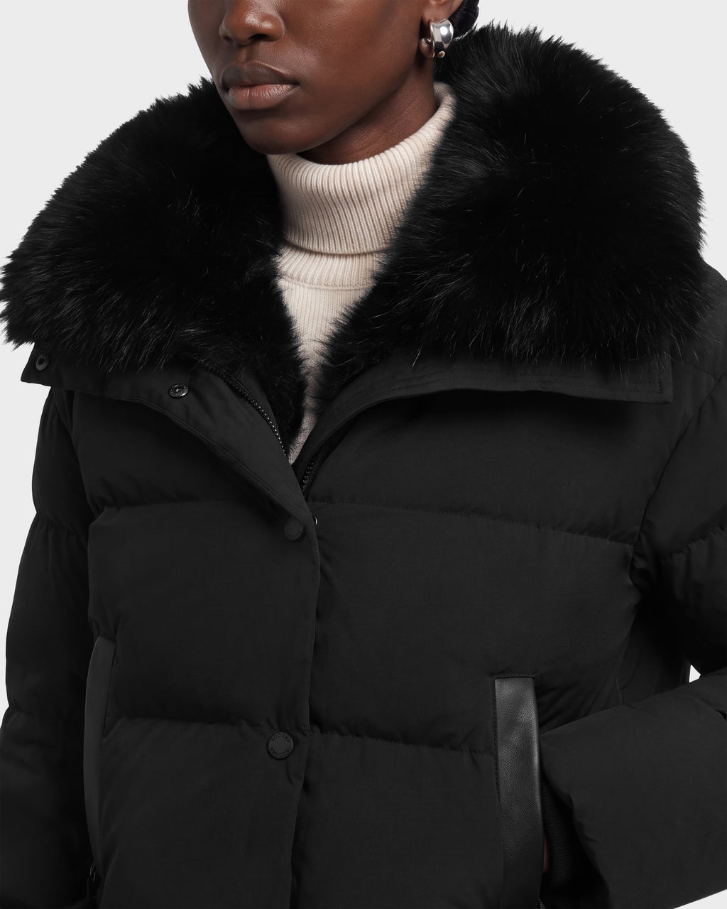 A person wearing a Rudsak ALONDRA - 8126992 Black puffer coat with a large fur collar over a cream-colored turtleneck.