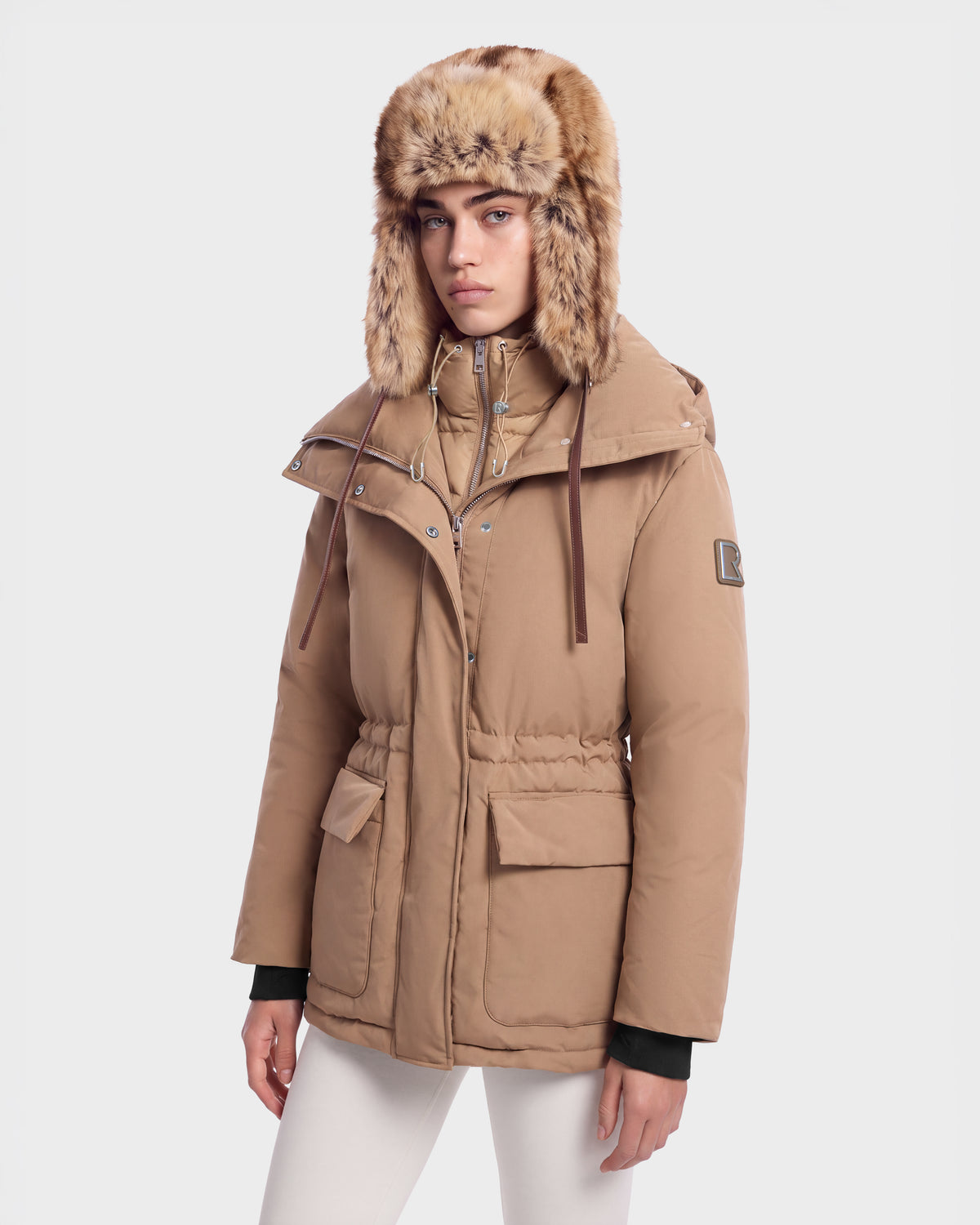 A person wears a camel-colored BHIMA - 8126515 Camel hooded parka with a faux fur trim and front pockets.