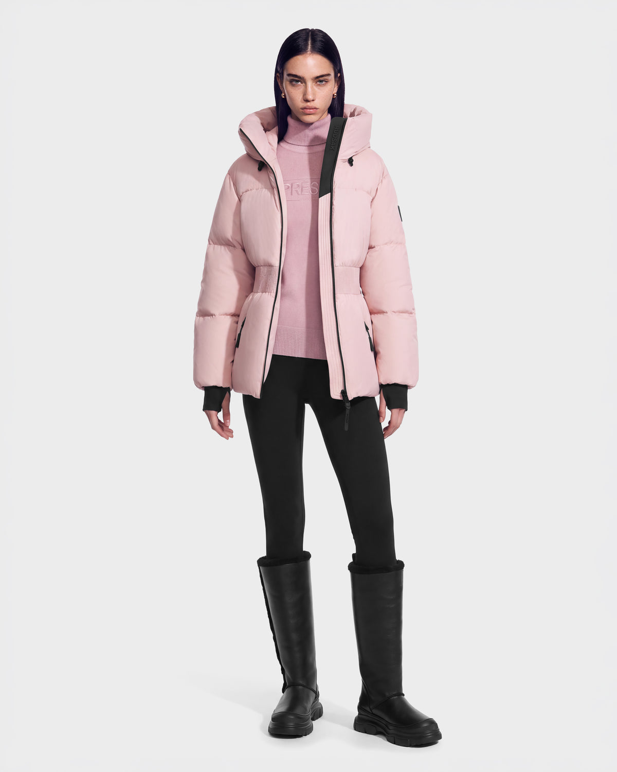 A model wearing a light pink DIAMOND - 8126772 Vintage Rose hooded, quilted puffer jacket over a matching turtleneck sweater, black leggings, and tall black boots.