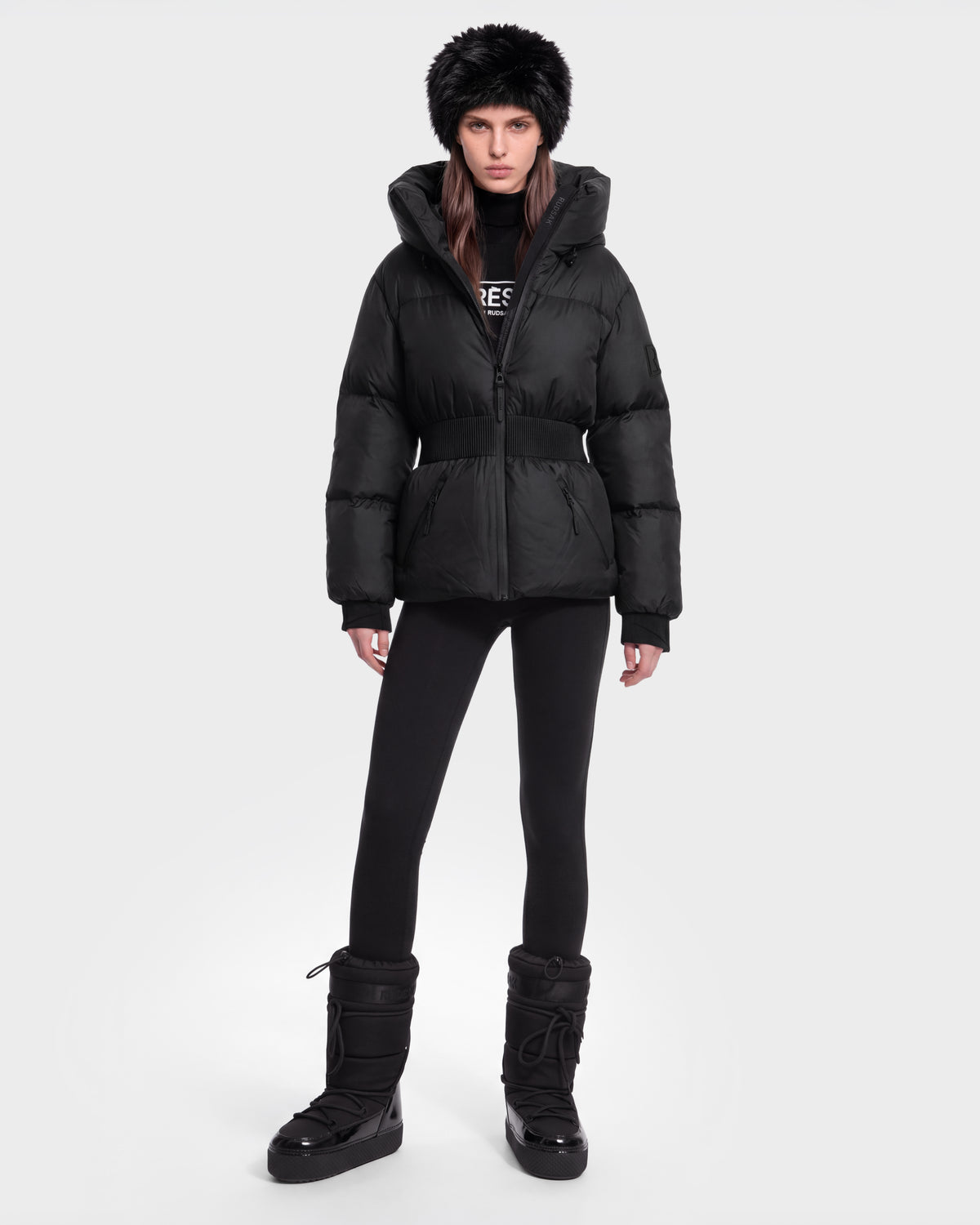A person wearing the Rudsak DIAMOND - 8126772 Black puffer jacket stands facing forward against a plain white background, paired with black leggings, winter boots, and a fur hat.