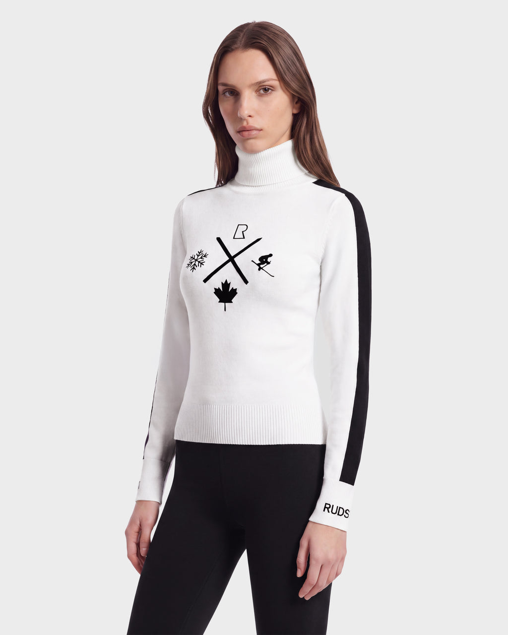 A woman wears the Rudsak ELIZABETH - 8524623 Optic White women’s ski turtleneck—a sleek white sweater featuring black winter sports icons and black side stripes—paired with black leggings, standing against a plain background. Elevate your winter wardrobe with this subtle après-ski style essential.