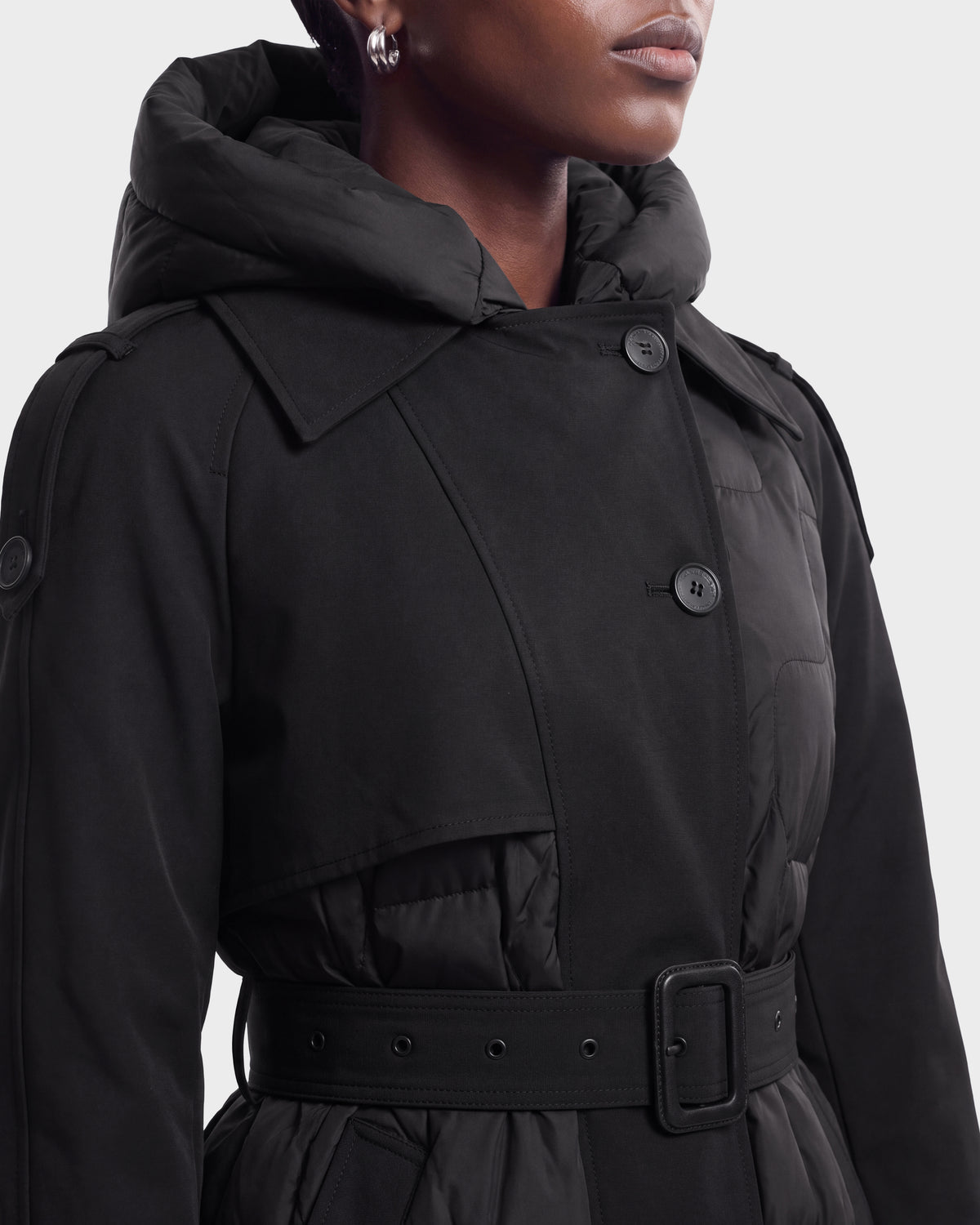A woman wearing the Rudsak ELVIE - 8126512 black belted coat with a hood and button details, shown from the shoulders down against a plain background.