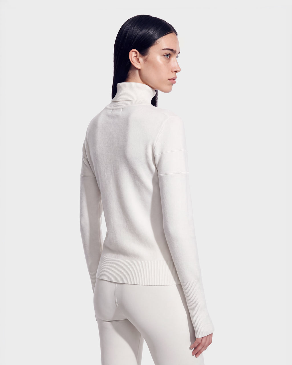 A person with long dark hair, wearing the ERIKA - 8525705 Iceland fitted white turtleneck sweater and matching white pants from Rudsak, stands facing away from the camera against a plain light gray background—a chic choice for any winter wardrobe.