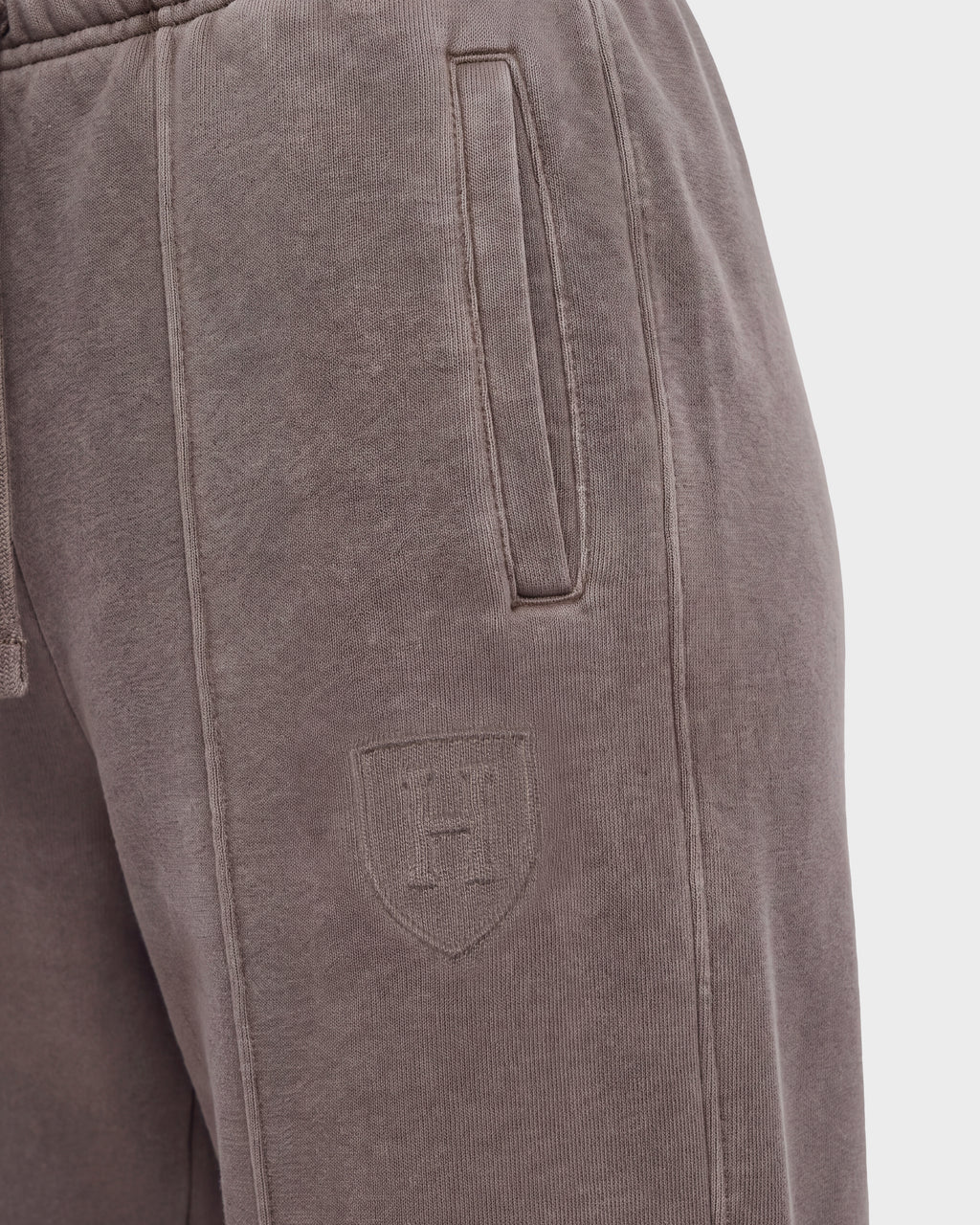 Close-up of the Rudsak GAELLE HV - H526902 Taupe heavyweight jersey jogger sweatpants in brown, featuring a slanted side pocket and a stitched shield emblem with the letter H on the upper thigh. The soft, slightly textured fabric evokes classic Harvard apparel style.