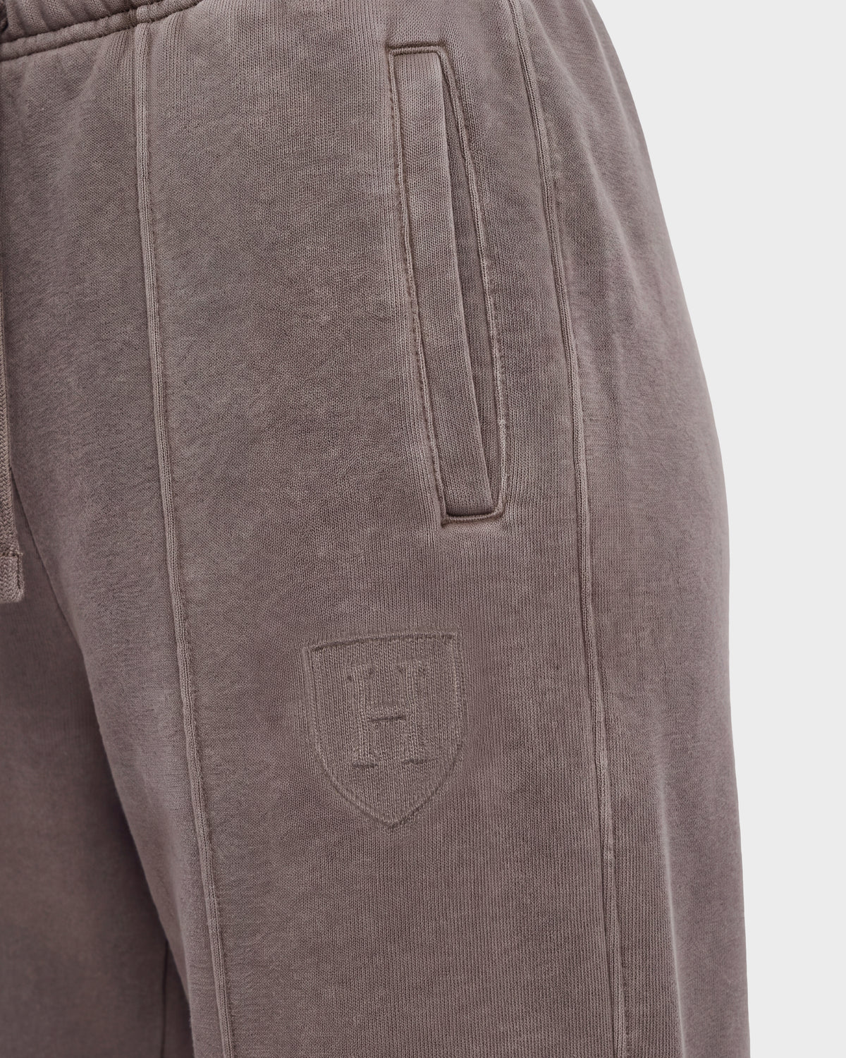 Close-up of the Rudsak GAELLE HV - H526902 Taupe heavyweight jersey jogger sweatpants in brown, featuring a slanted side pocket and a stitched shield emblem with the letter H on the upper thigh. The soft, slightly textured fabric evokes classic Harvard apparel style.