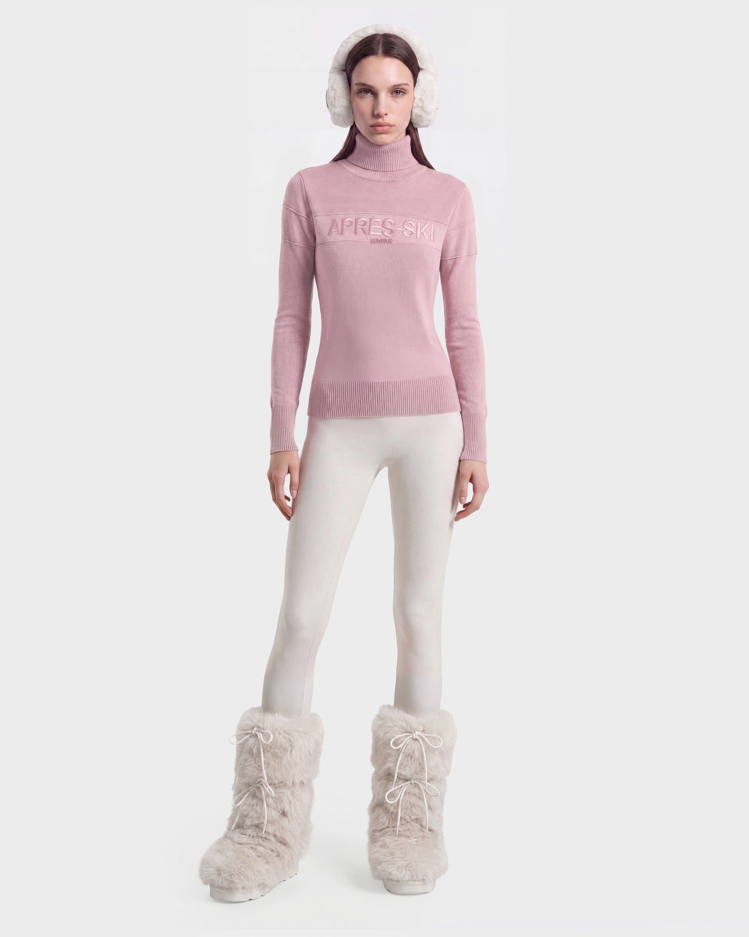 A woman stands against a plain light background, showcasing an effortlessly chic winter look in the Rudsak ERIKA - 8525705 Vintage Rose turtleneck sweater, styled with white leggings, fluffy white earmuffs, and large furry white snow boots—an ensemble that refreshes the seasonal wardrobe.