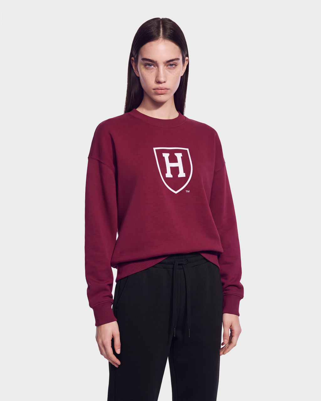 A person with straight brown hair wears the Rudsak HARVARD ONE CRIMSON SWEATSHIRT 9524507—a maroon unisex sweater adorned with a white shield and H logo—paired with black pants and standing against a plain gray background, capturing classic Ivy League style.