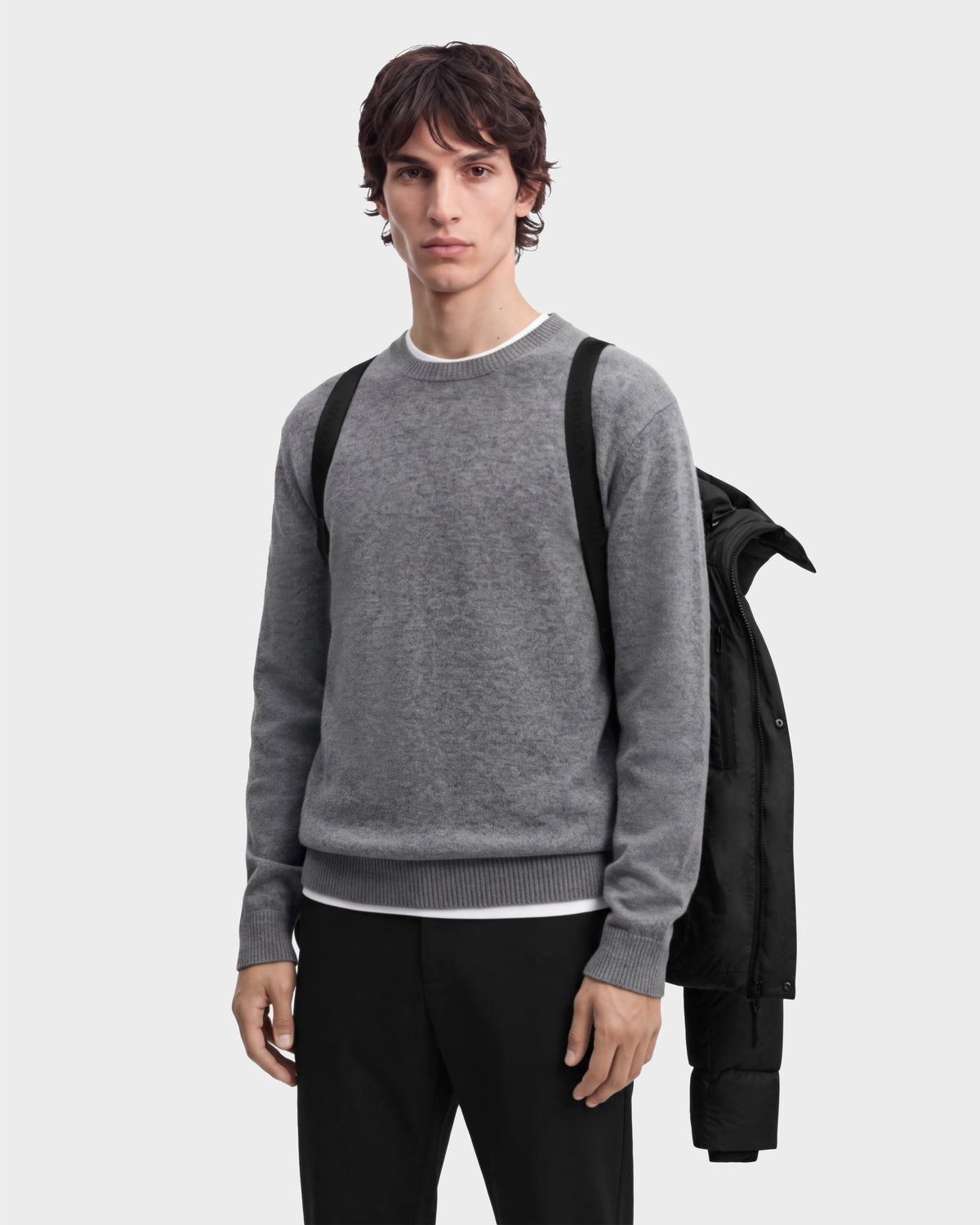 A man wears a grey crew-neck sweater over a white t-shirt, with a JASON - 6126803 Black jacket hanging off one shoulder.