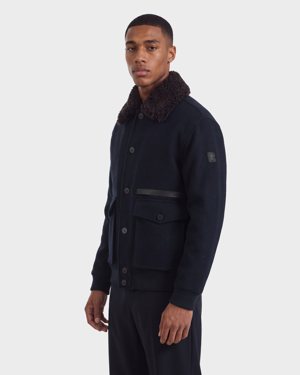 A man wears a navy blue wool-blend Rudsak JULIUS - 6126723 Navy bomber jacket featuring a dark faux-fur collar and two large front pockets.