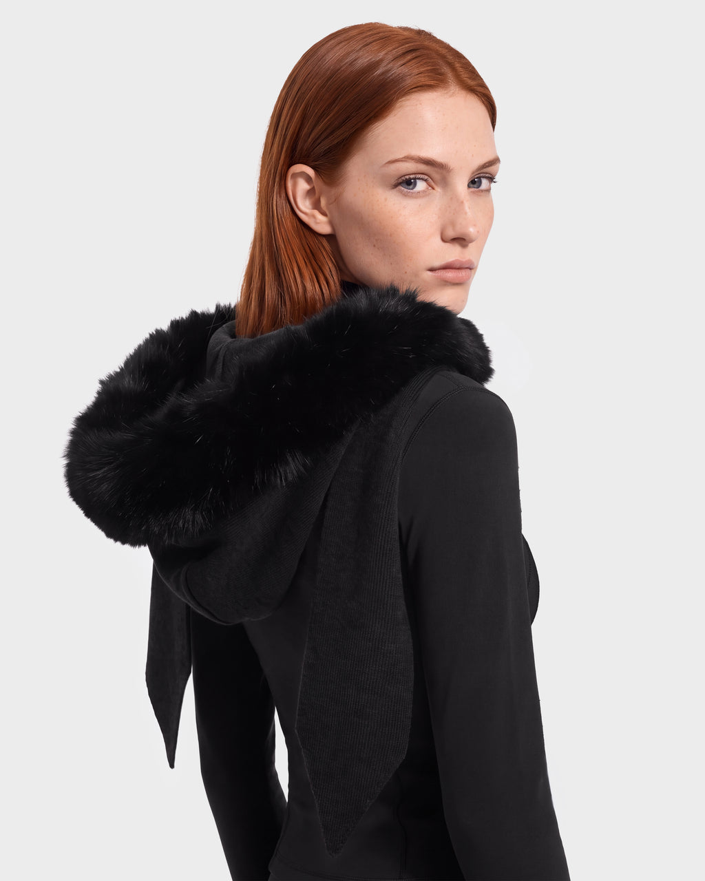 A woman with straight red hair wears the Rudsak KARYMA - 8426507 Black hood-and-scarf hybrid, featuring a plush black fur trim, as she looks over her shoulder against a plain light background.