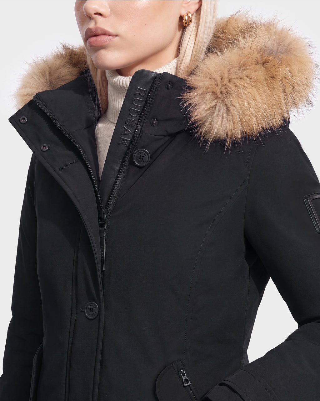 A woman wears the Rudsak KEYSHIA - 8126517 Black down parka with a fur-lined hood over a cream turtleneck, showing only the lower half of her face and upper body.
