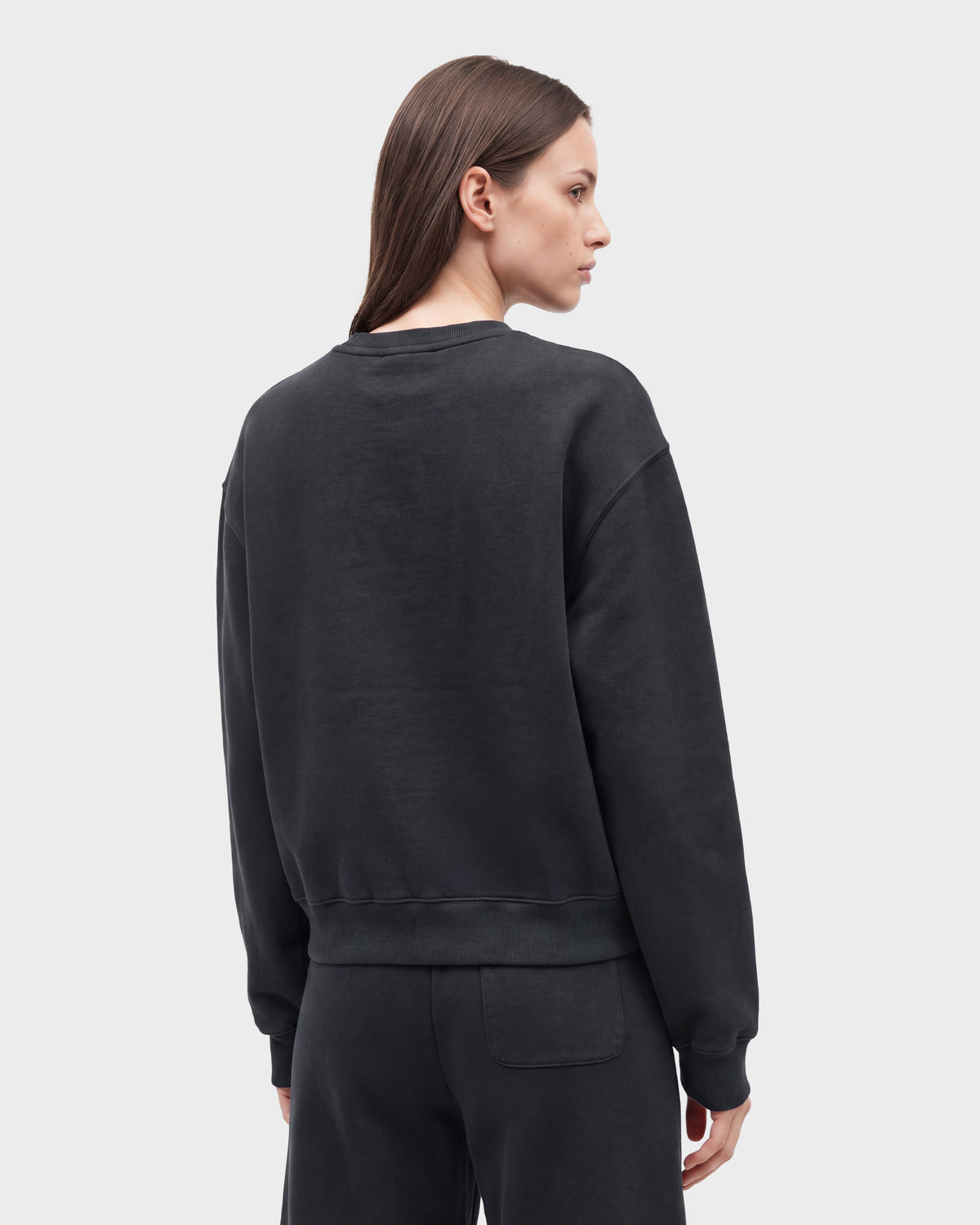 A woman with straight brown hair is shown from the back wearing the Rudsak LEXYE HV - H526900 Titanium loose-fitting black sweatshirt and matching black pants against a plain light gray background.