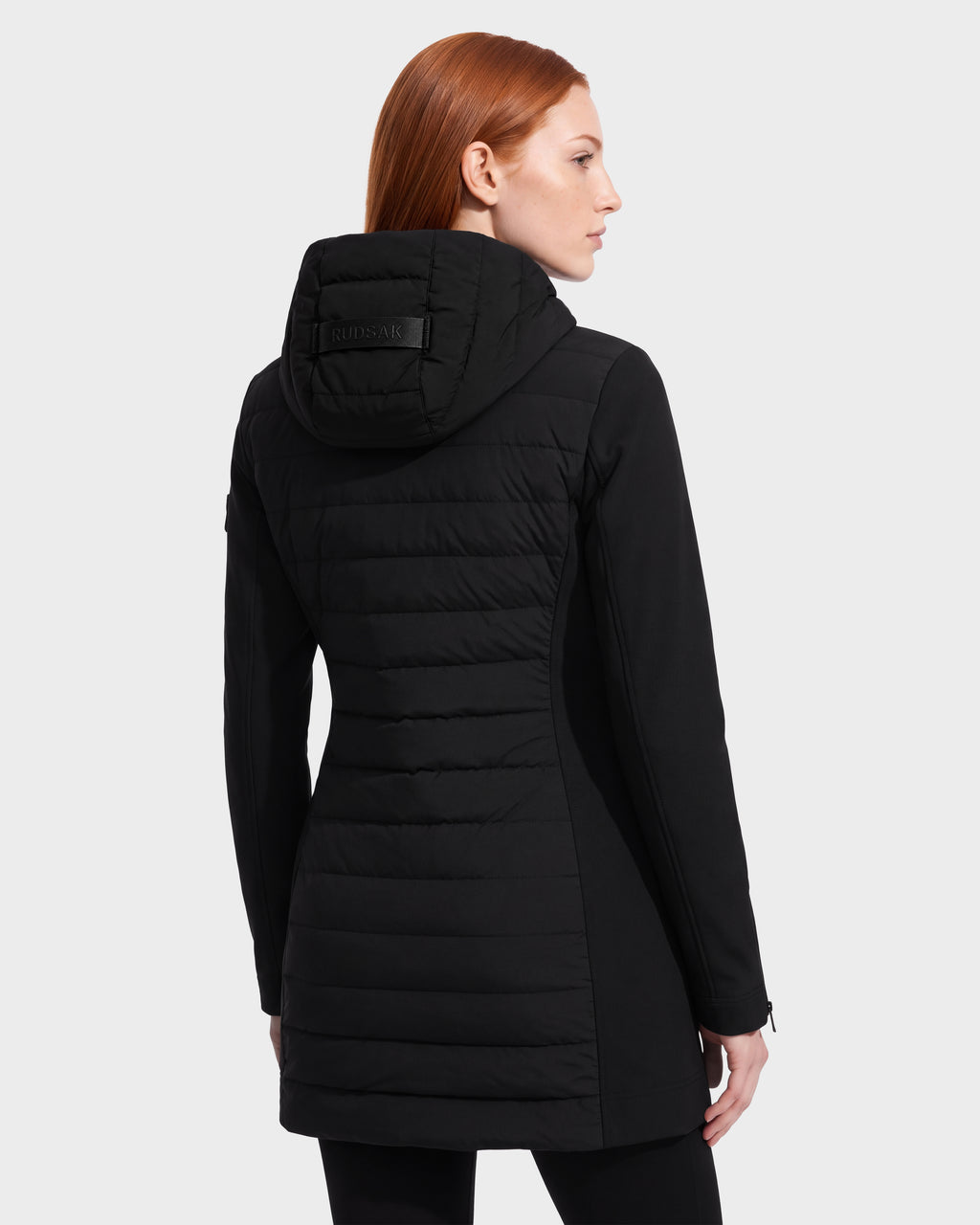 A woman with straight red hair is shown from the back, standing against a plain white background, wearing the Rudsak MERINA - 8125056 Black women’s long hooded winter coat inspired by light jackets.