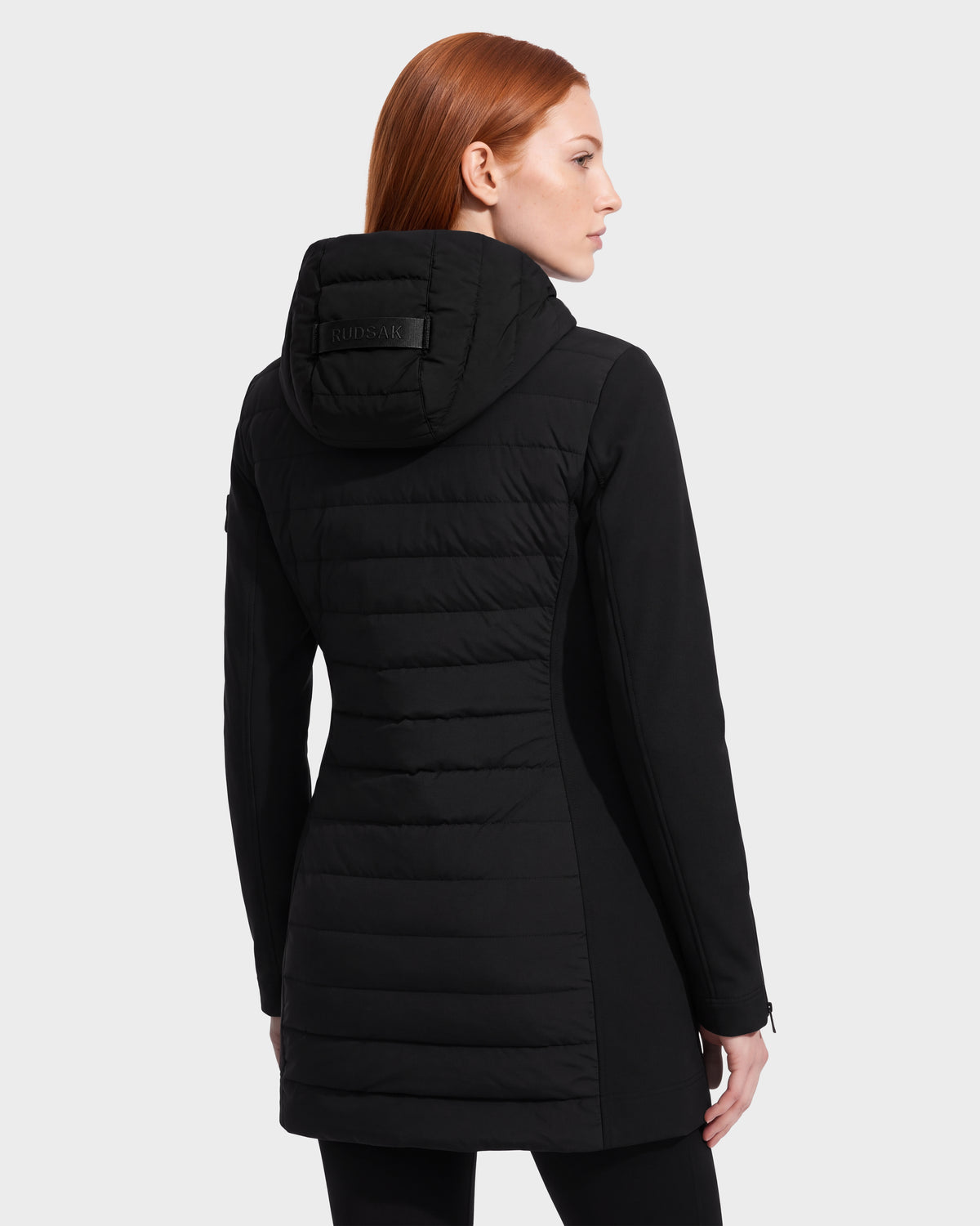 A woman with straight red hair is shown from the back, standing against a plain white background, wearing the Rudsak MERINA - 8125056 Black women’s long hooded winter coat inspired by light jackets.