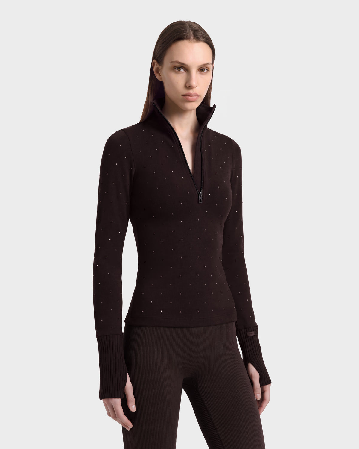 A woman wearing the NEVA - 8526524 Espresso, a dark brown après-ski top with a quarter-zip, scattered silver studs, and ribbed cuffs with thumbholes.