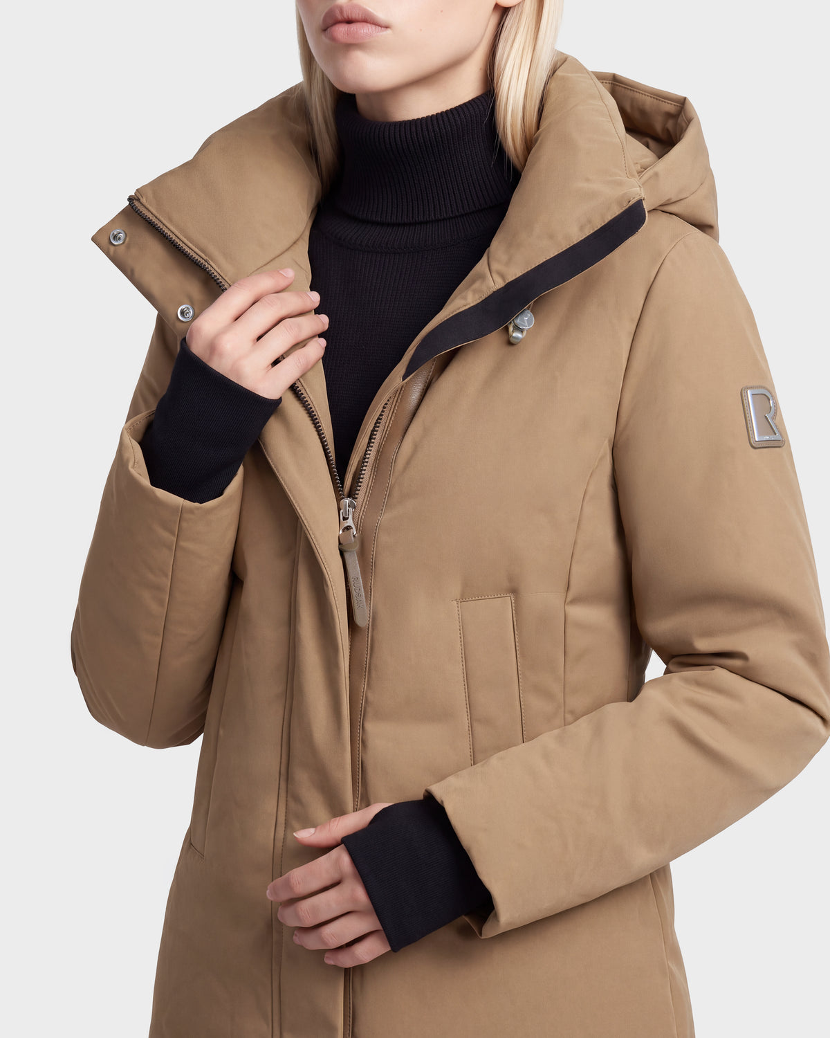 A woman wears the Rudsak OPAL - 8126618 Camel parka over a black turtleneck. The coat features a sleeve patch, snaps on the collar, and RDS-certified down fill. Her hand rests near the zipper.
