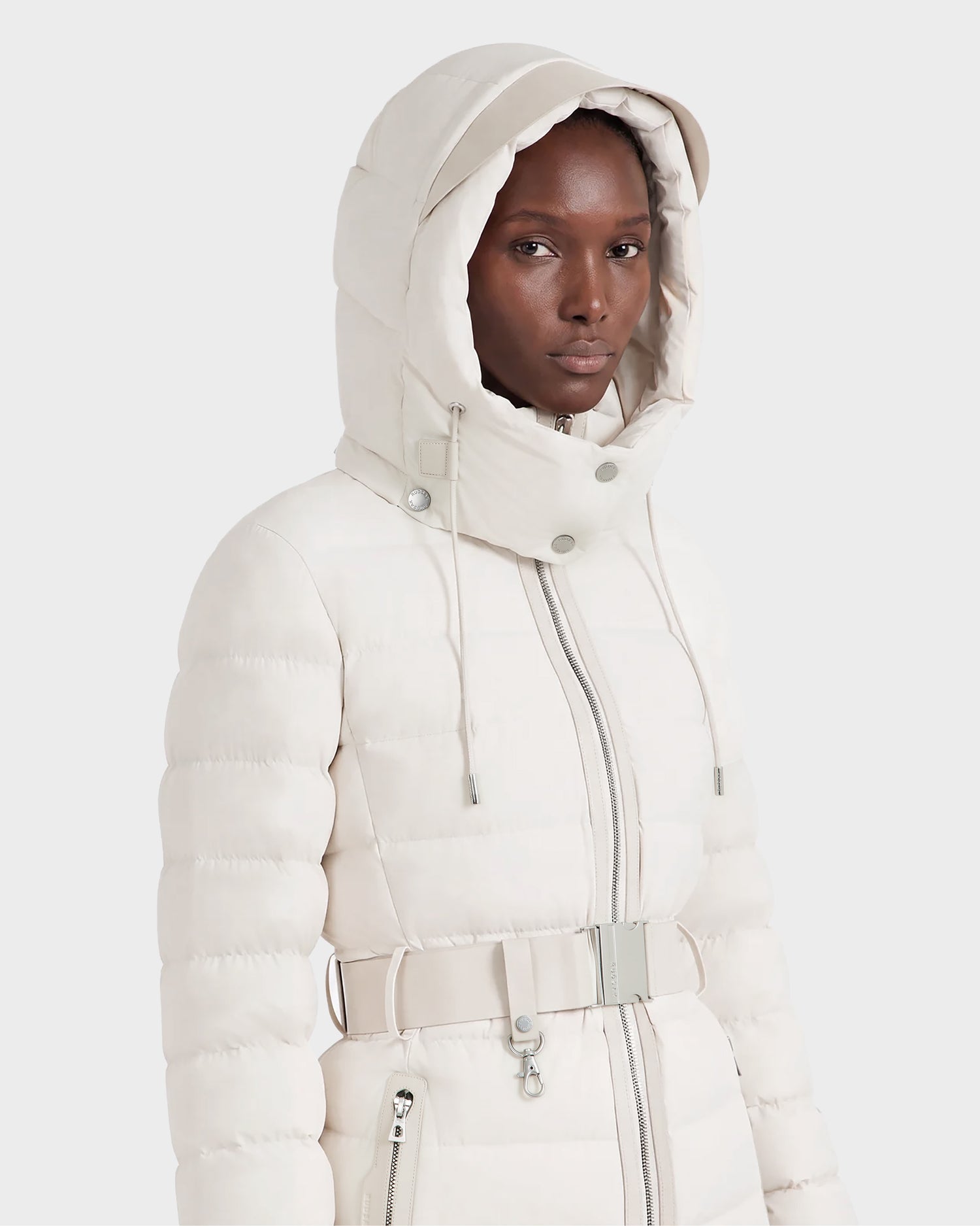 A person stands against a plain background wearing the Rudsak PELIN S - 8124813 Vanilla, a light beige women’s down puffer jacket featuring a high collar, front zipper, belted waist, and zippered pockets.