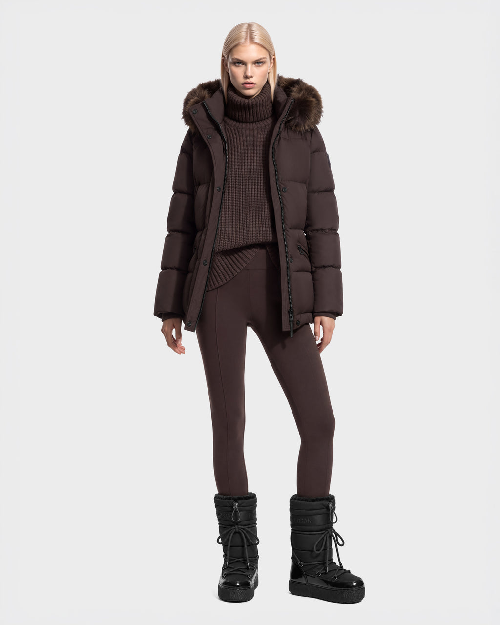 A person wearing a brown Rudsak GRACE FF - 8126535 Espresso hooded puffer jacket with faux fur trim, a matching ribbed turtleneck sweater, leggings, and black winter boots.