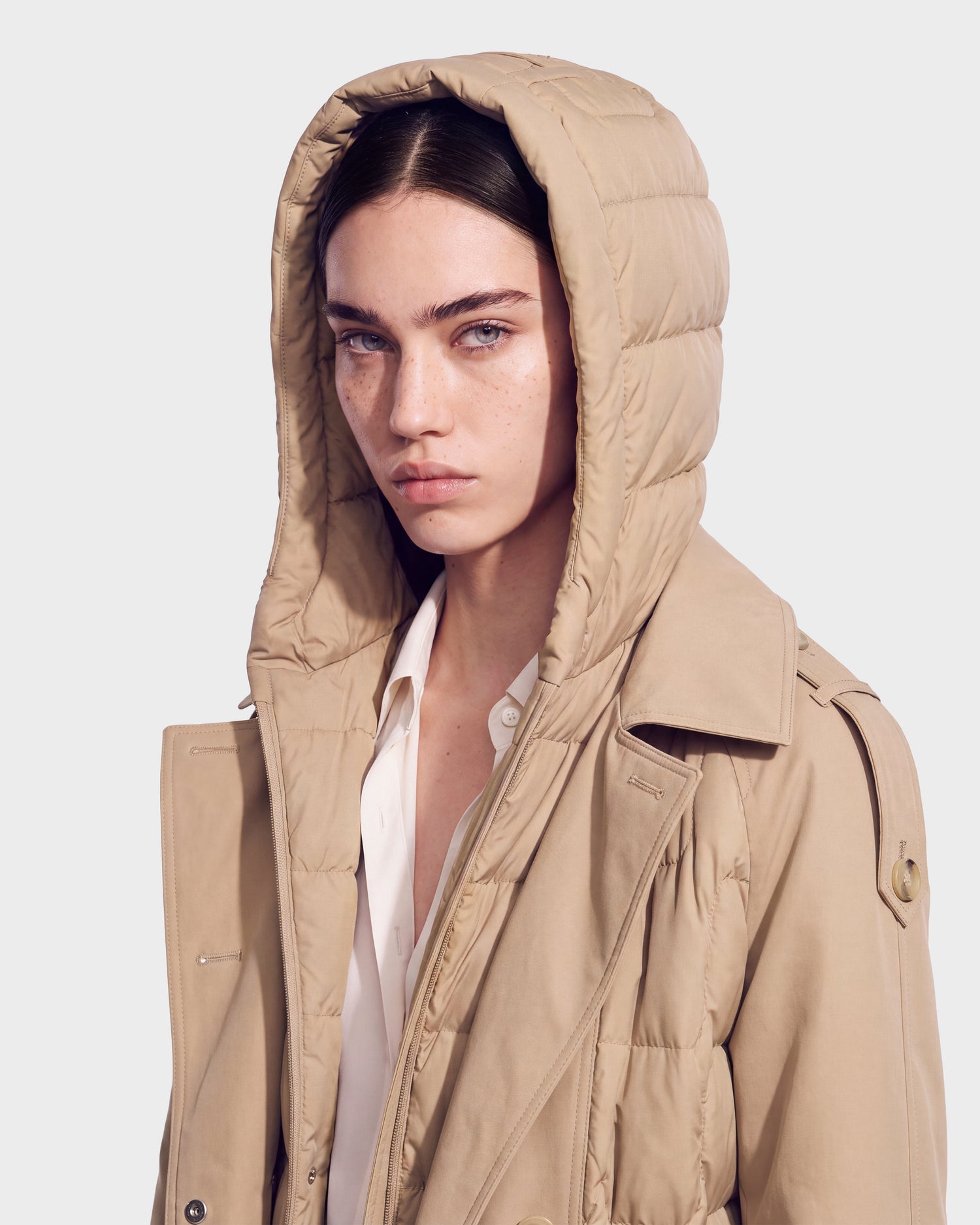 A woman with light eyes and brown hair wears the ELVIE - 8126512 Almond women’s winter coat by Rudsak, styled like a down trench coat, over a white shirt. She looks directly at the camera with a serious expression against a plain background.