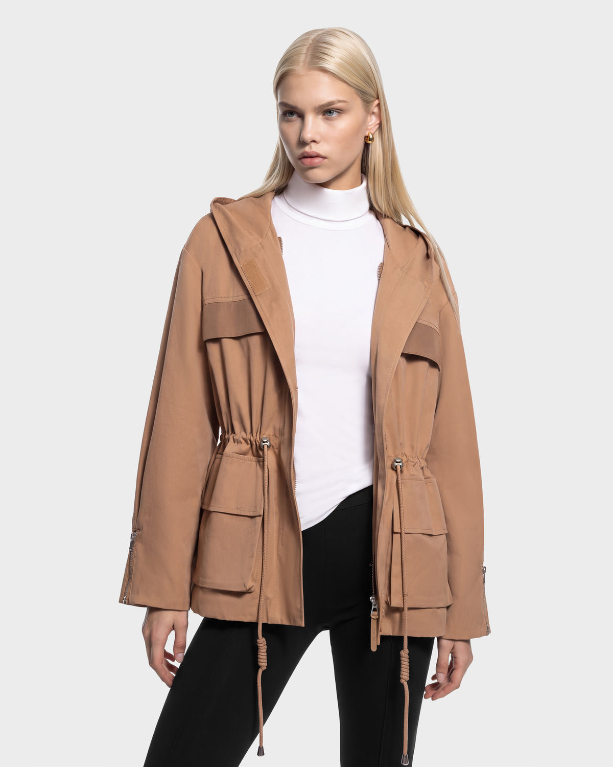 A woman wears a camel-colored ROSALINDA - 8126523 Camel hooded jacket with a drawstring waist and flap pockets over a white turtleneck and black leggings.