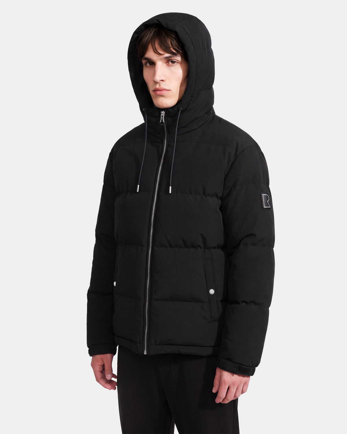 A young man stands facing forward against a plain white background, wearing the Rudsak STERLING EL - 6126759 Black puffer jacket featuring a hood, zipper closure, and front pockets.