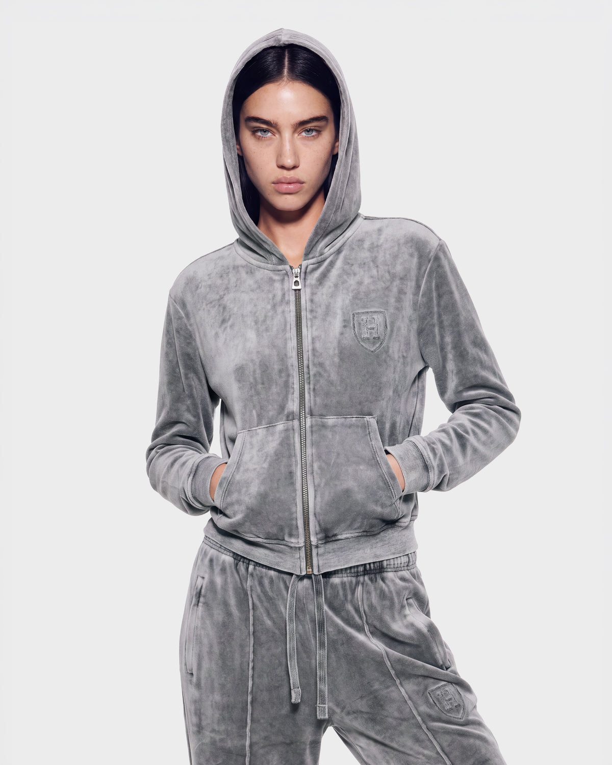 A person stands with hands in pockets, looking straight ahead against a plain background, wearing the Rudsak TYRA HV - H526904 Titanium velour tracksuit featuring a zip-up hoodie and drawstring pants.