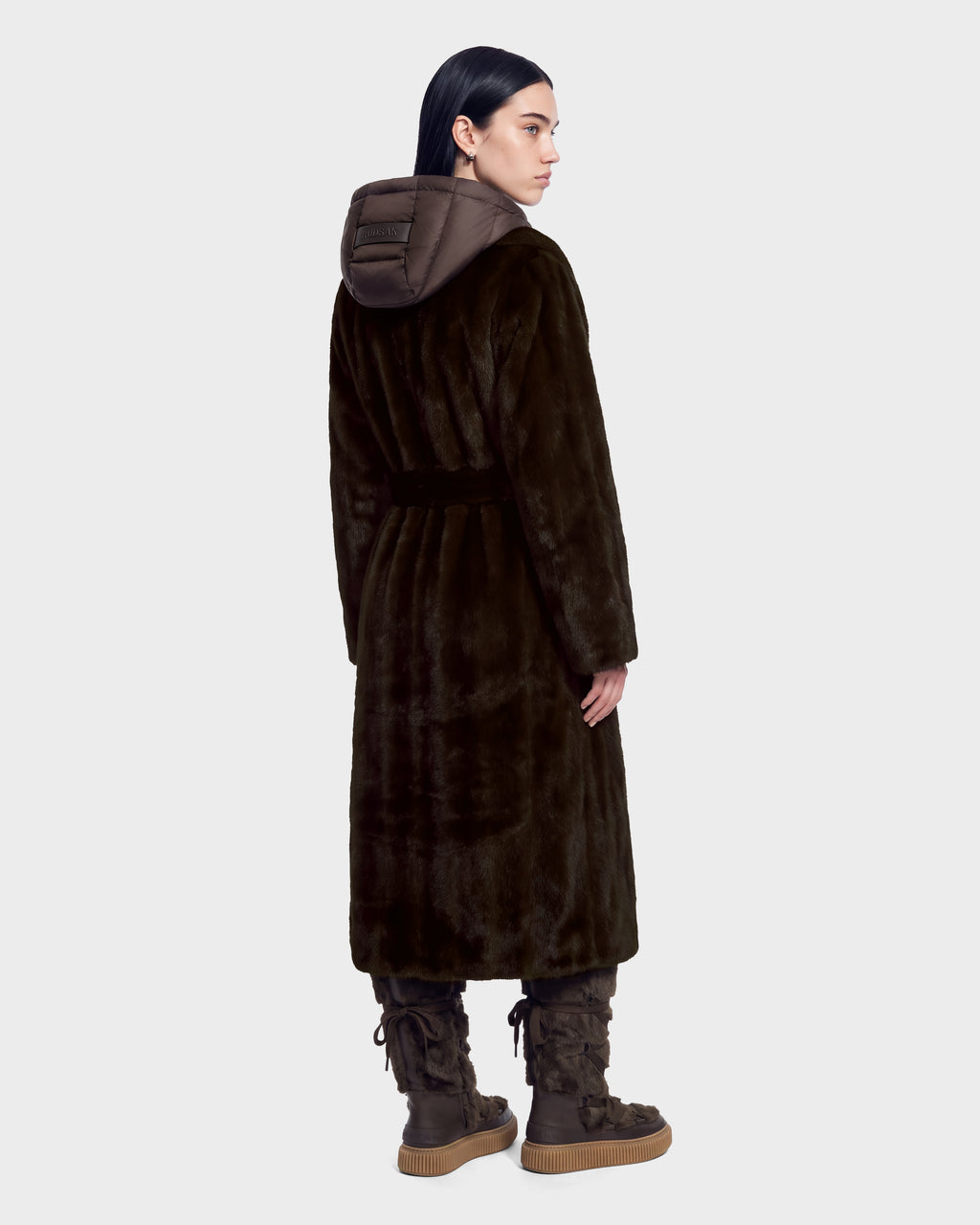 A person seen from behind wears the Rudsak VIVIENNE - 8126528 Espresso dark brown faux-fur long coat with a hooded collar and matching boots with thick, lugged soles, embodying chic winter city dressing.
