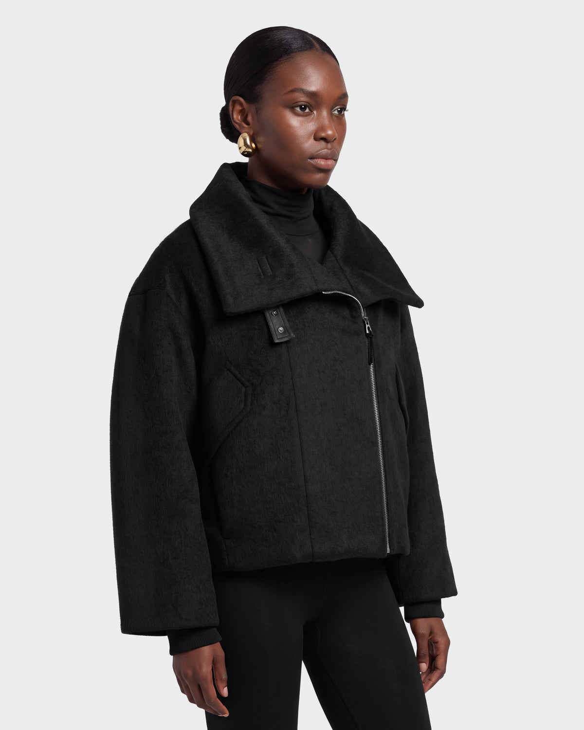 A woman stands against a plain light background, looking to the side with a neutral expression. She is wearing the ZARIA - 8126999 Black oversized jacket by Rudsak, paired with gold hoop earrings and black pants.