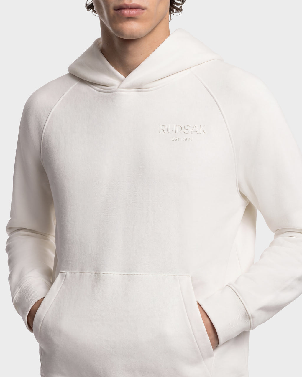 A person wearing a cream-colored Rudsak BRIAR - 9525001 Vanilla M unisex hoodie with a front kangaroo pocket and the RUDSAK EST. 1994 logo embossed on the chest.